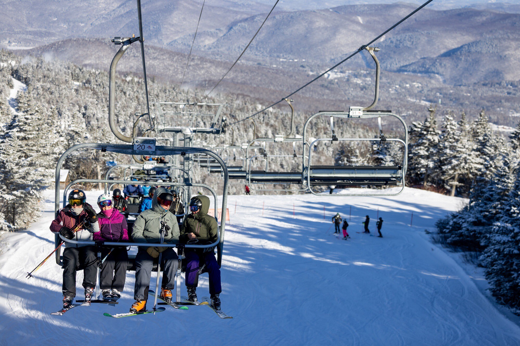 Skiers on a chairlift with snowy slopes and trees in the background.