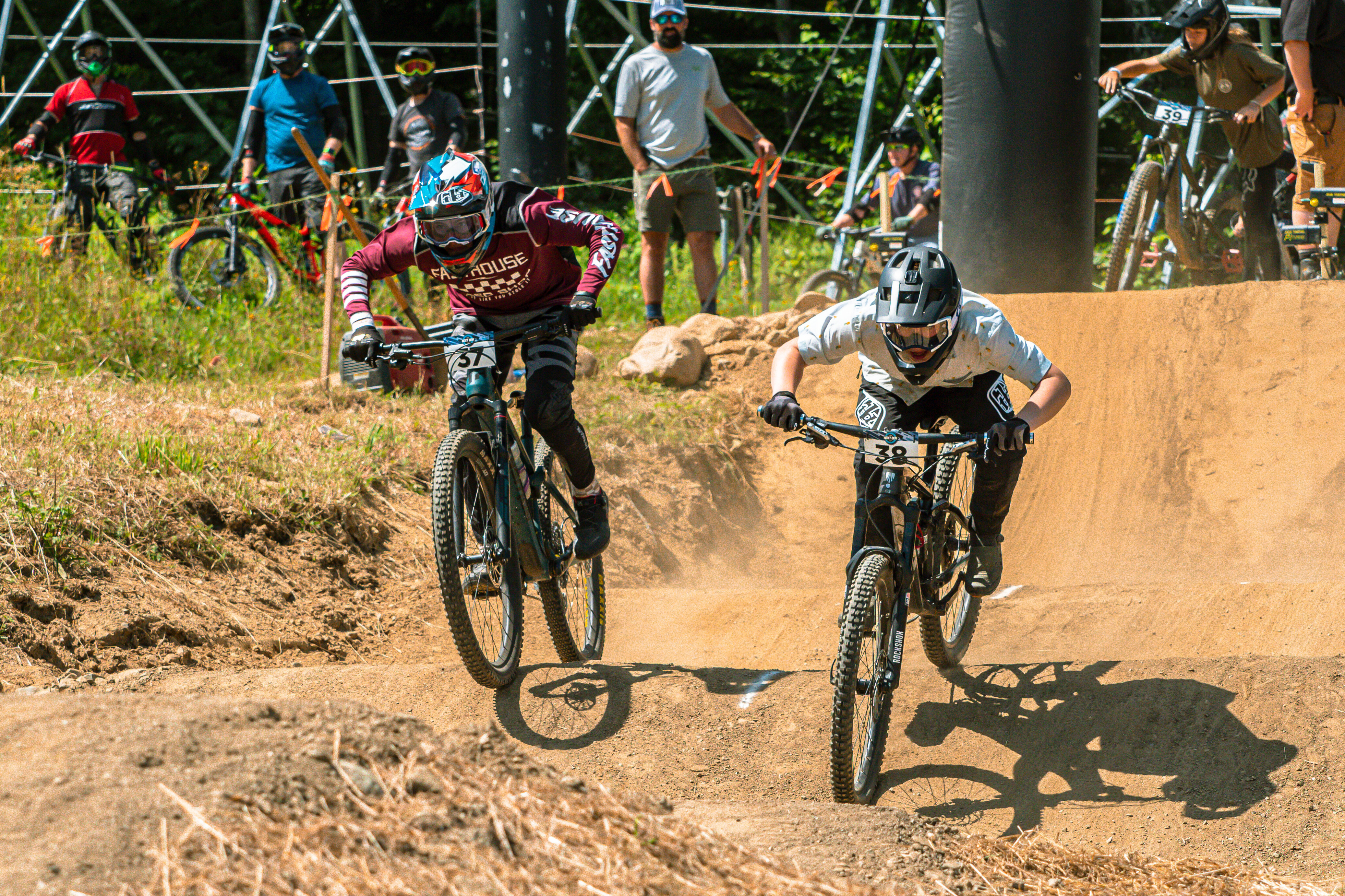 Two mountain bikers race down a dirt track with spectators in the background.