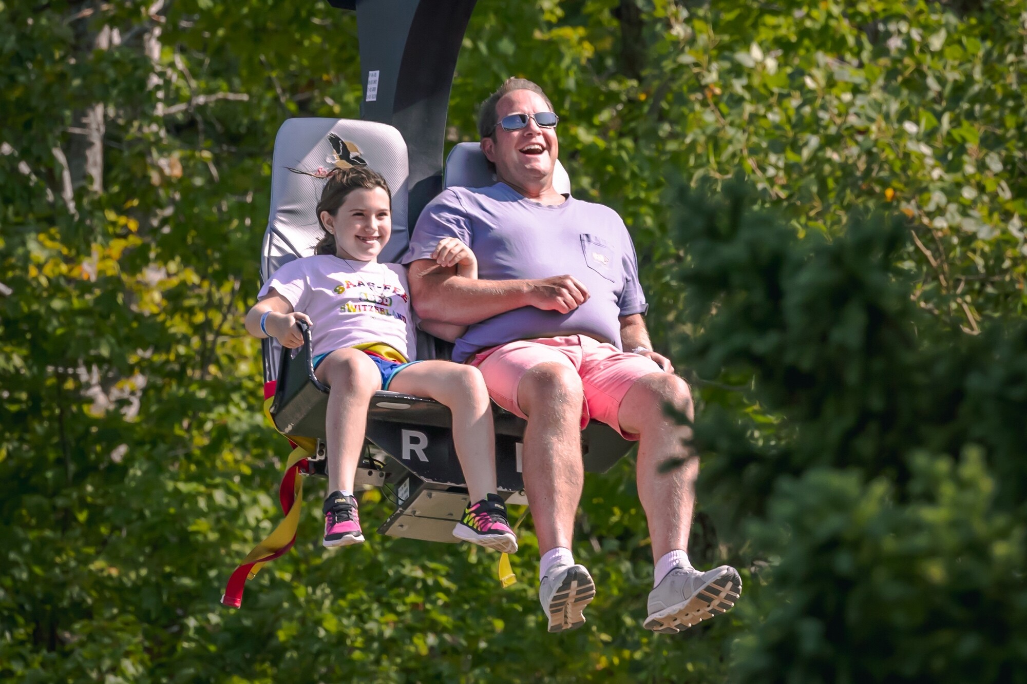A man and a chile smile joyfully while riding a chairlift amidst green trees at the Killington Mountain Resort Adventure Center