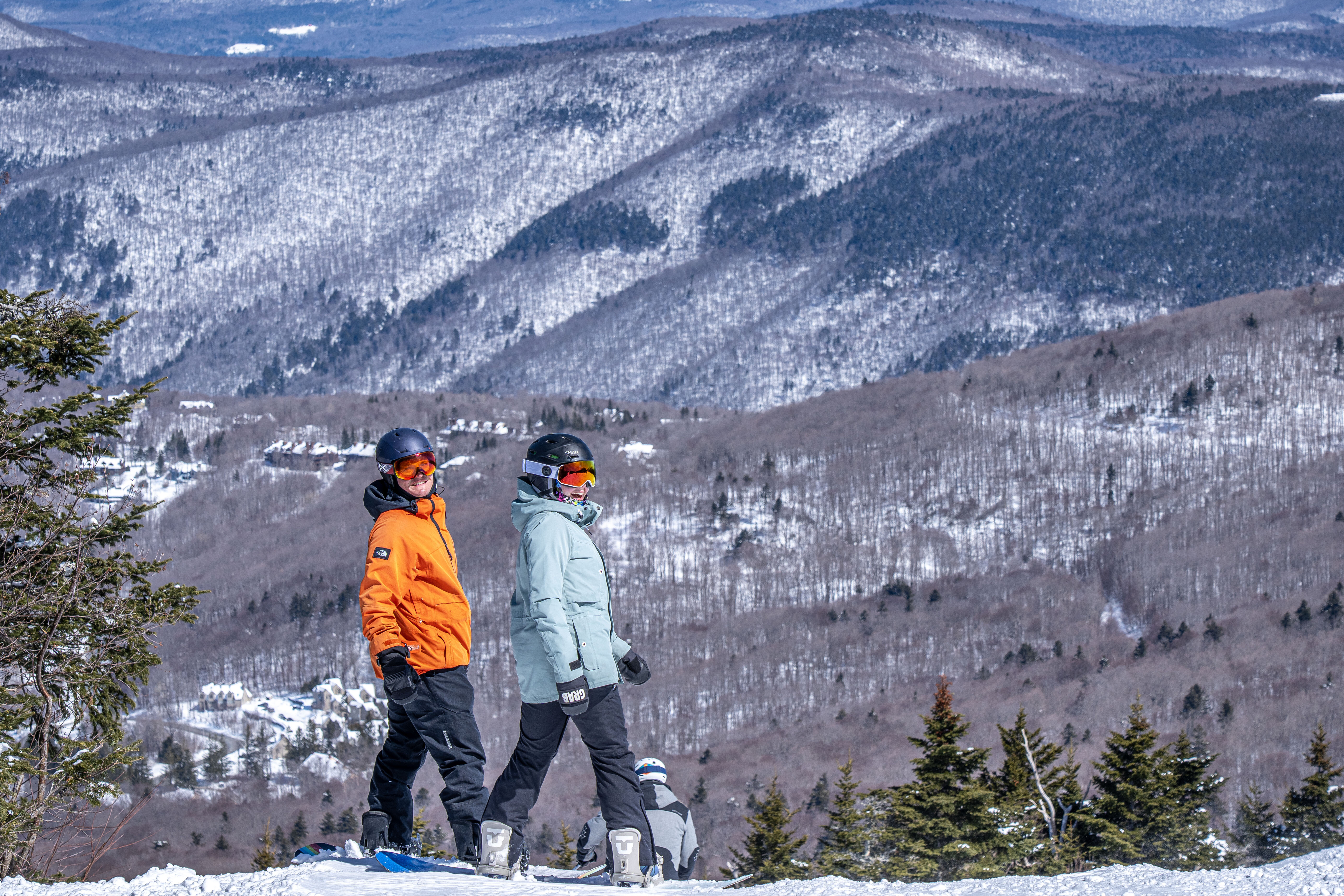 Two skiers pose on a snowy mountain with scenic forested hills in the background.