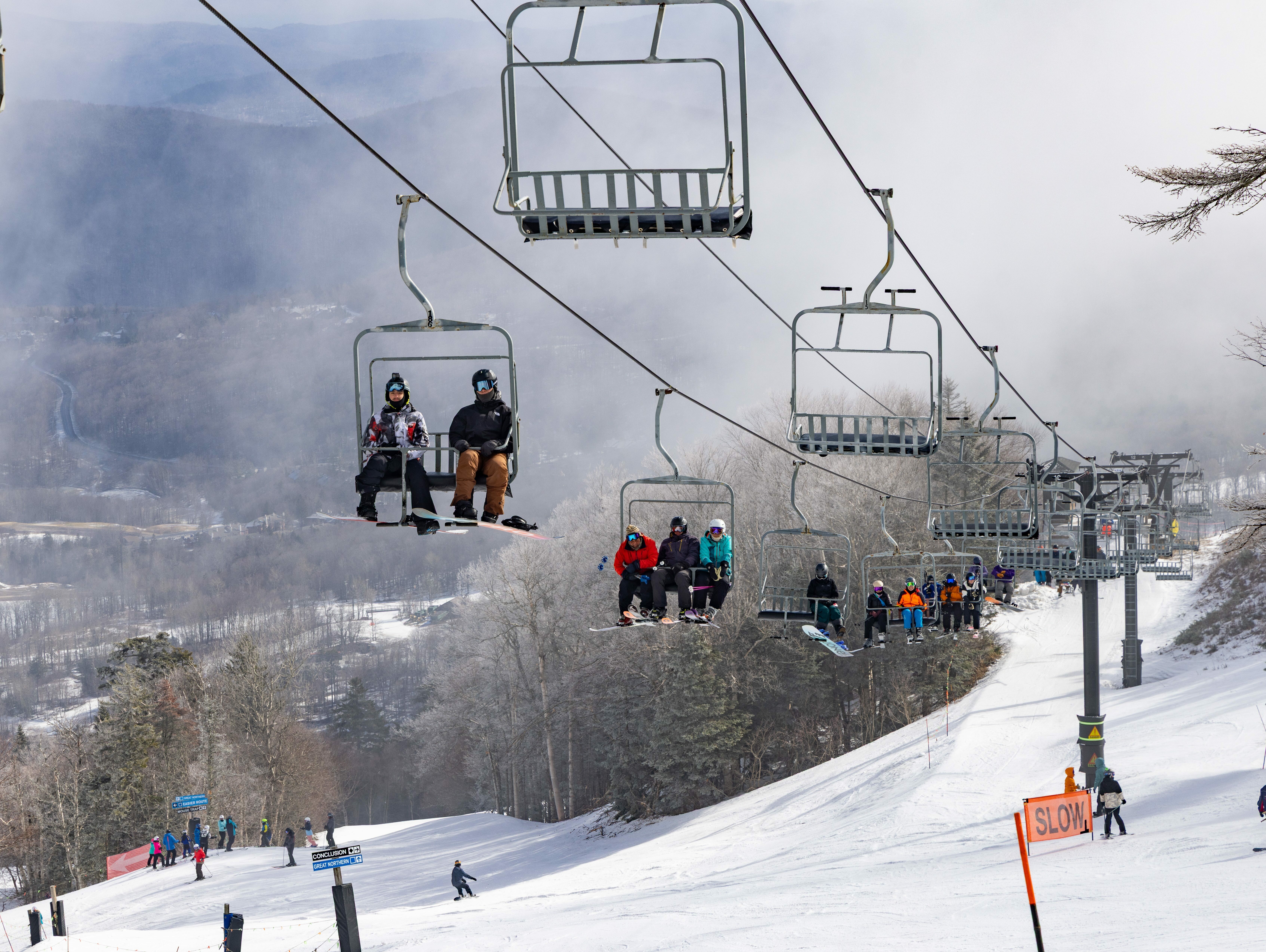Skiers on a chairlift above a snowy slope with trees in the background.