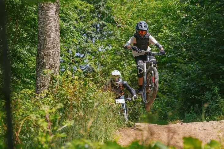Two mountain bikers navigate a forest trail, one jumping off a small ramp.