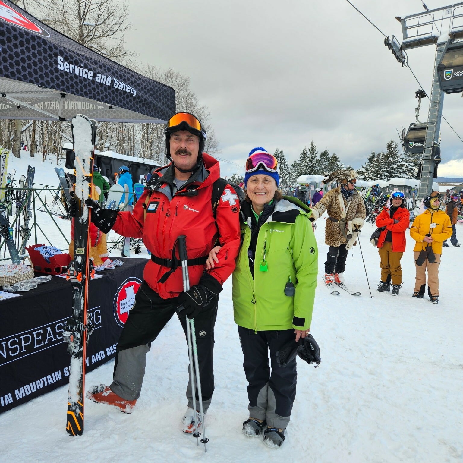 Two skiers pose together in winter gear at a ski resort, with a snow-covered backdrop.