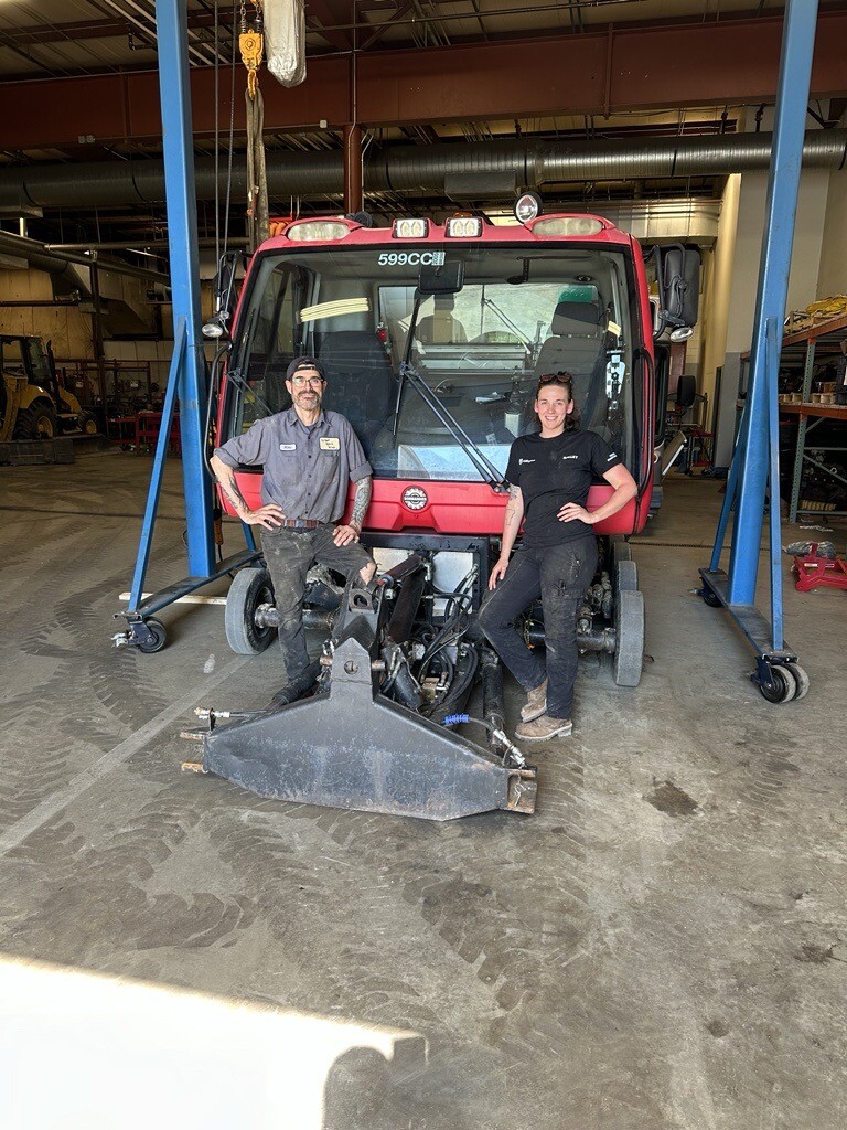 Two mechanics stand beside a red vehicle in a service shop, with equipment visible.