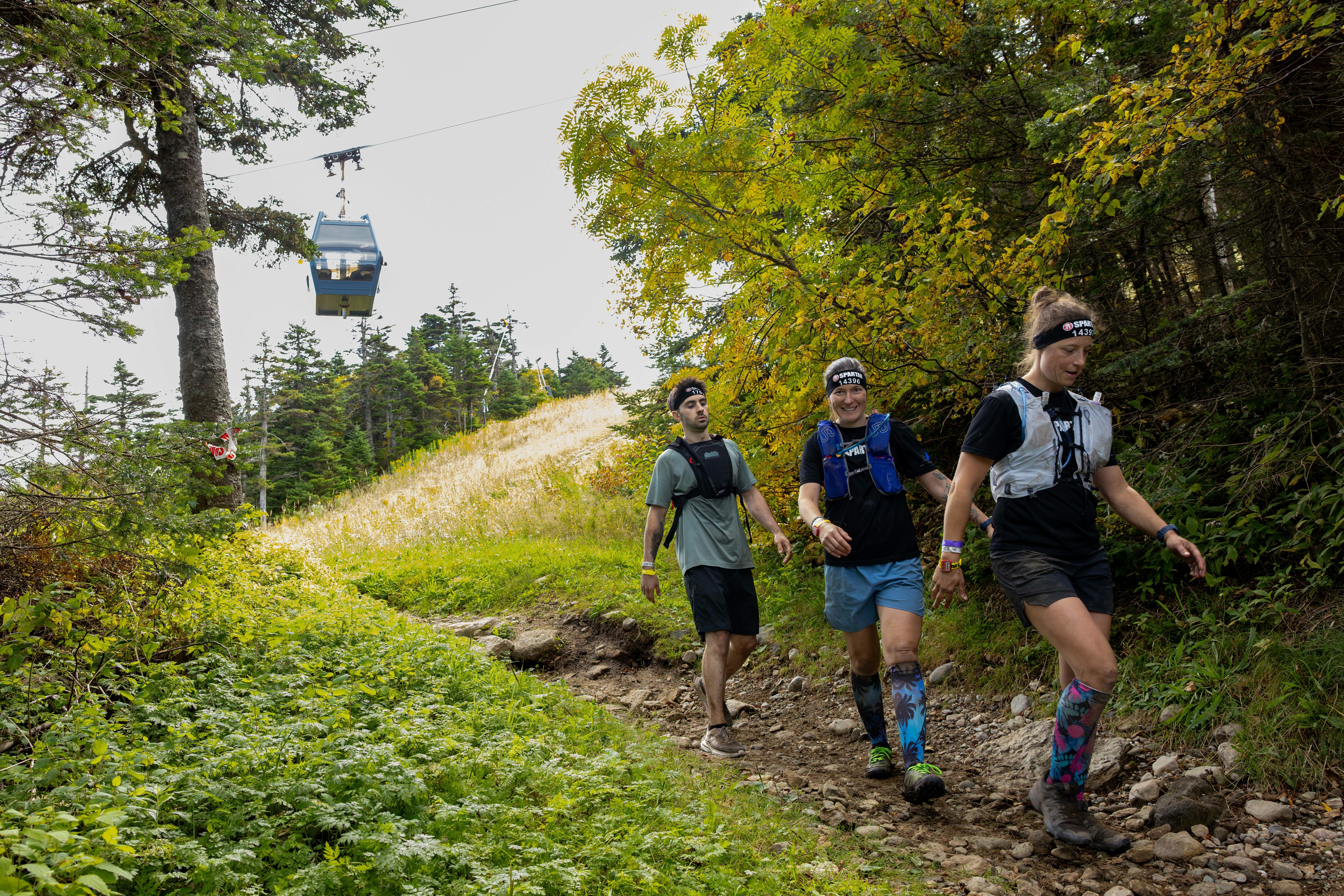 Three hikers on a trail with a gondola overhead, surrounded by greenery and trees.