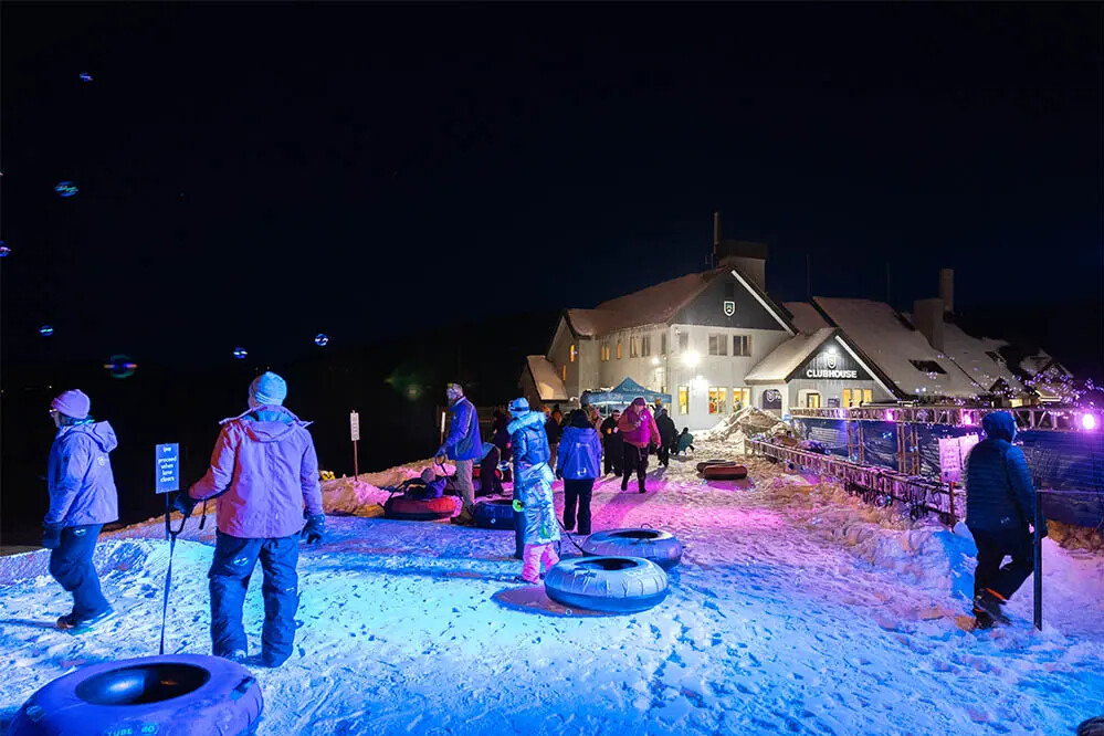 Snowy night scene with people tubing near a clubhouse, illuminated by colorful lights.