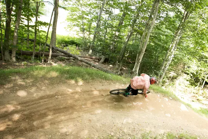 cyclist in a pink shirt leans into a turn on a dirt bike trail through trees.