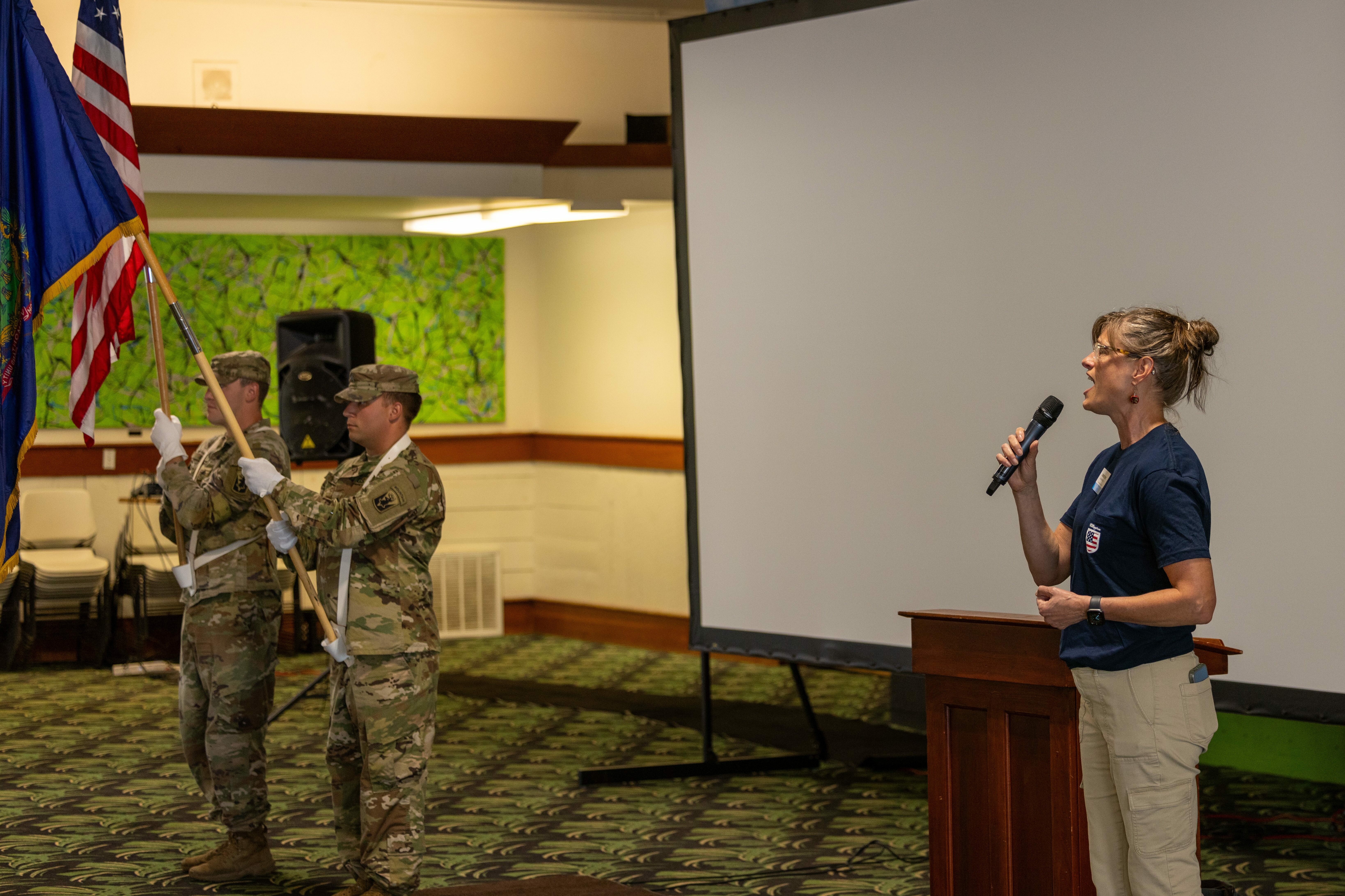 A woman sings into a microphone while soldiers hold flags in a meeting space.