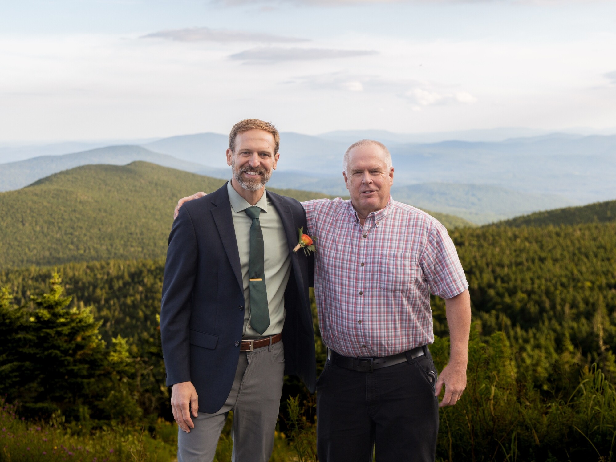 Two men pose together with mountains in the background, smiling at the camera.
