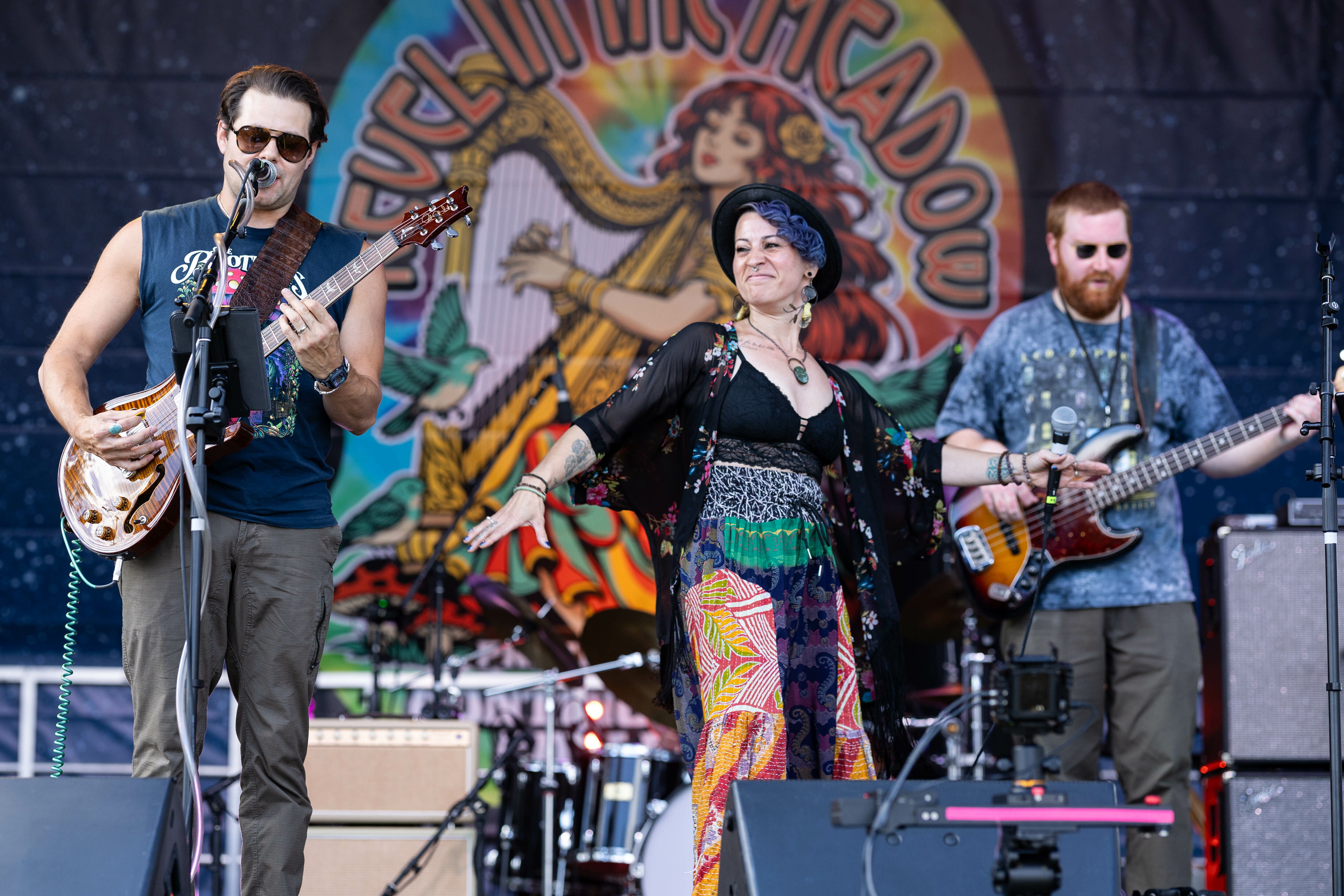 A band performs on stage, featuring a guitarist, singer in colorful attire, and a bassist.