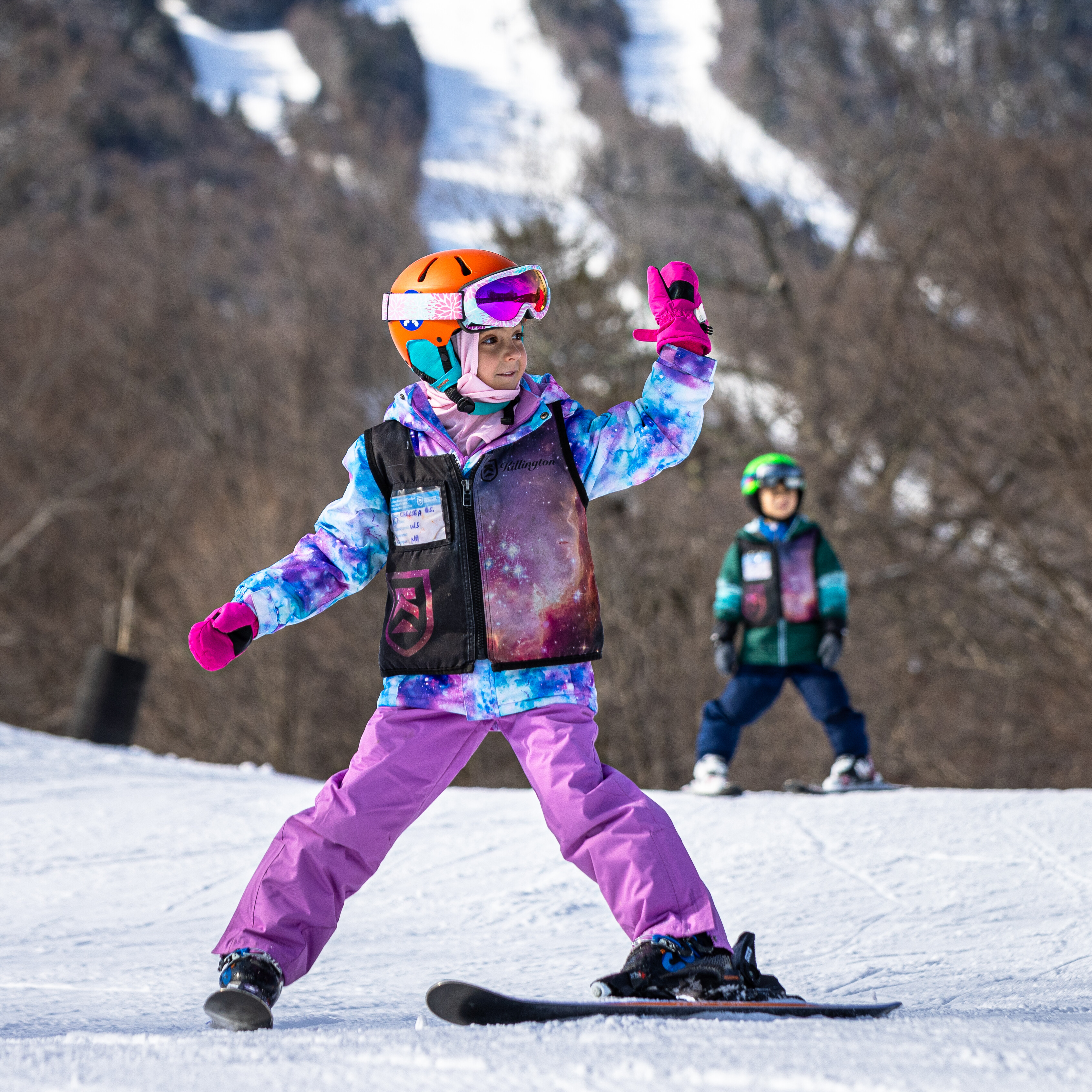 A girl snowboards on a slope, joyfully posing, with a boy in the background.