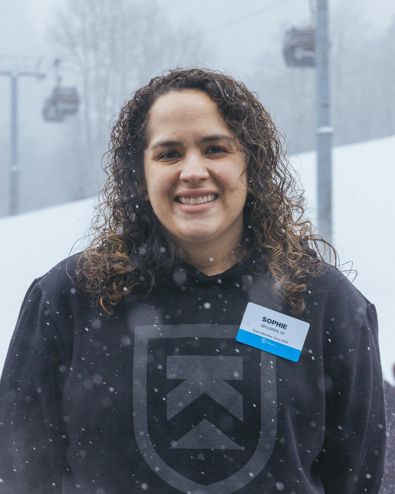 Smiling woman wearing a hoodie and name tag, snowy ski resort backdrop.