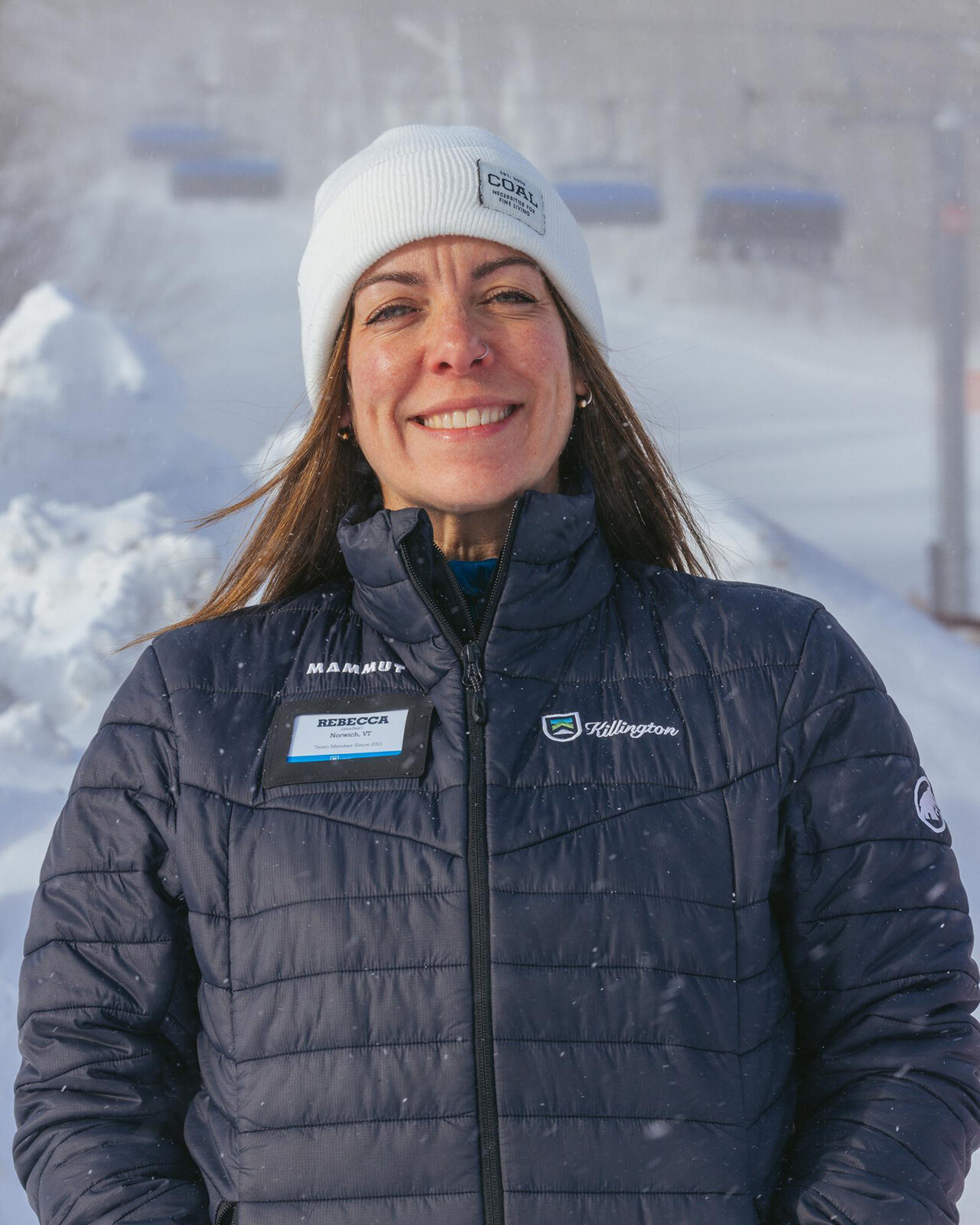 Woman in a winter jacket and beanie, smiling in a snowy landscape.