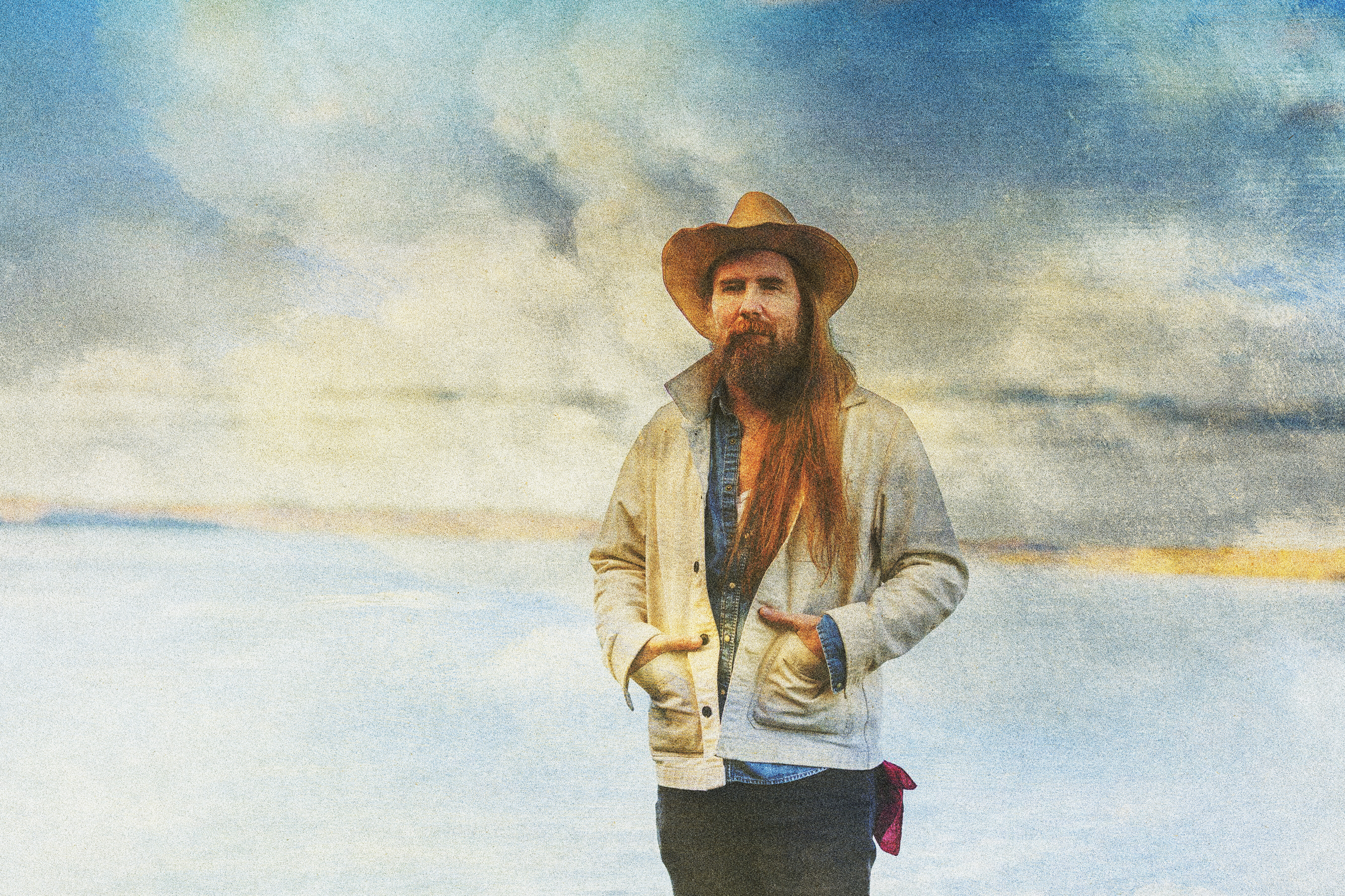 Bearded man in a cowboy hat stands by water under a colorful sky.