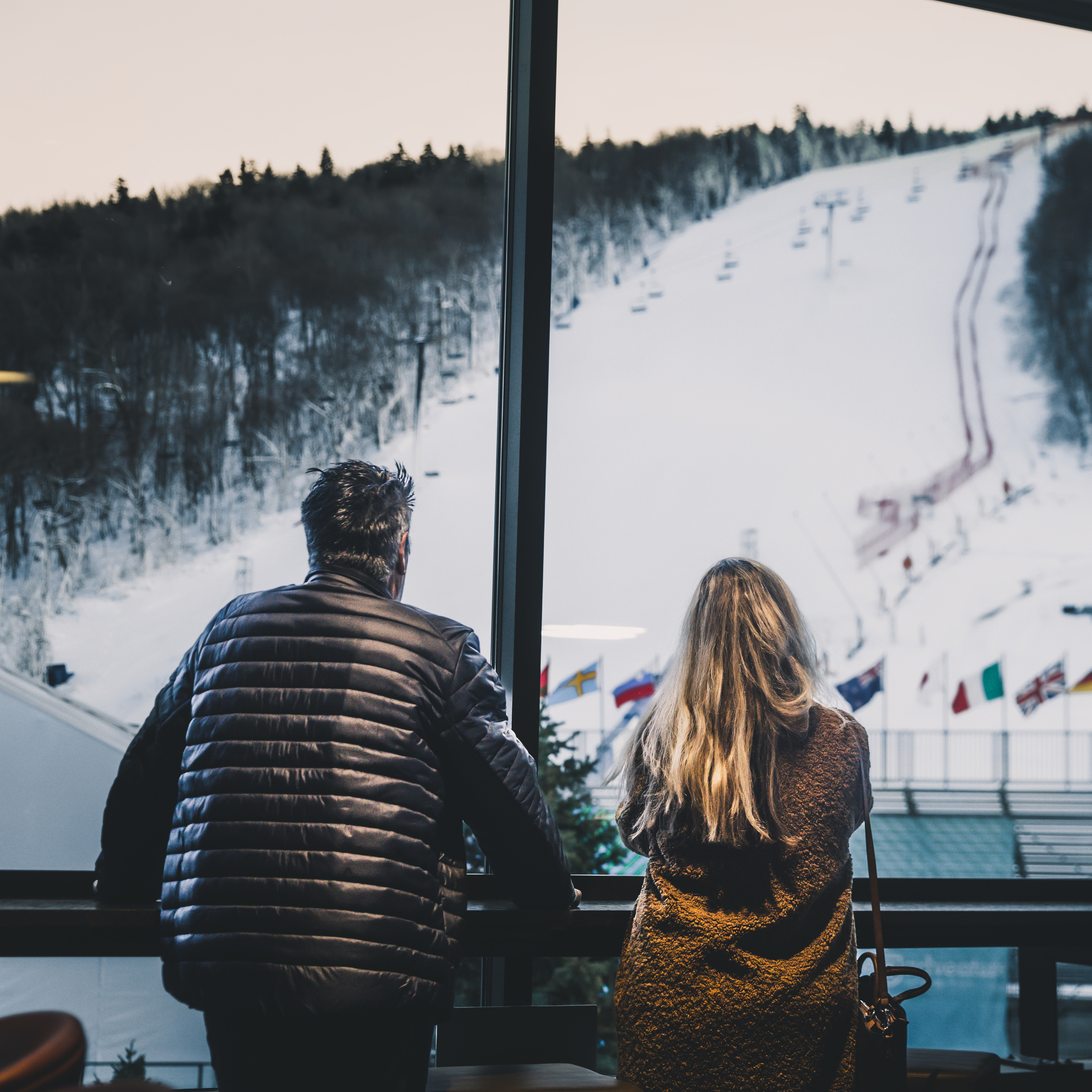 A couple gazes out the window at a snowy ski slope and flags.