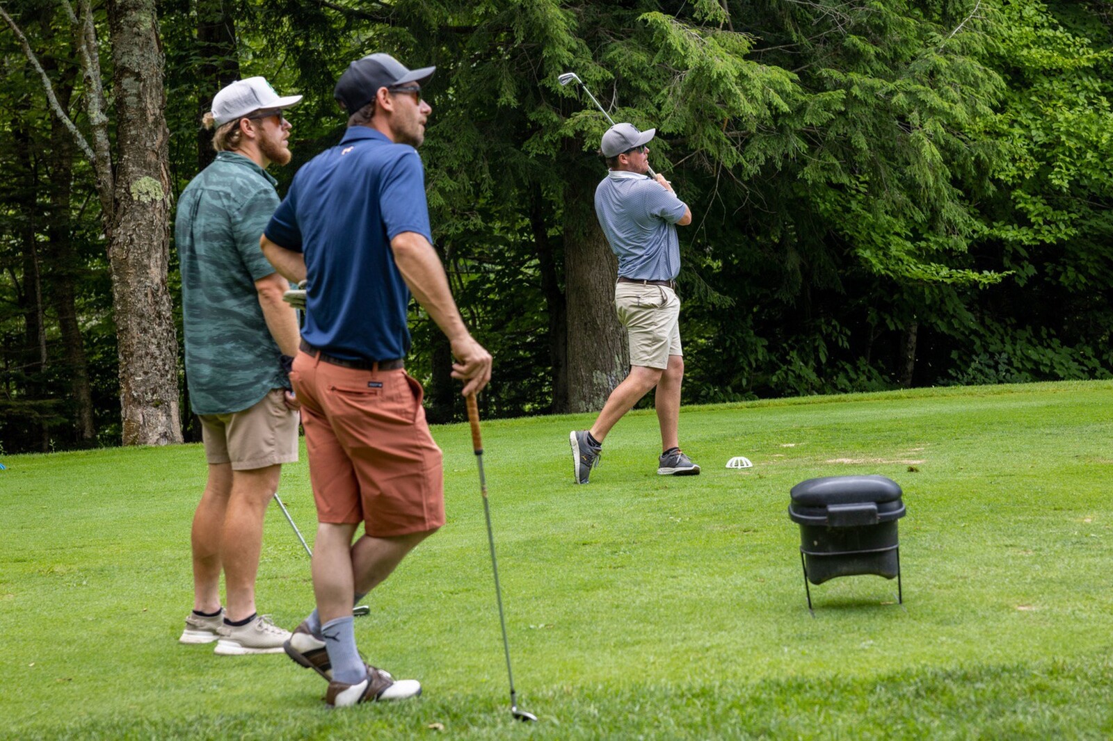 Three men on a golf course; one swings a club while the others watch.