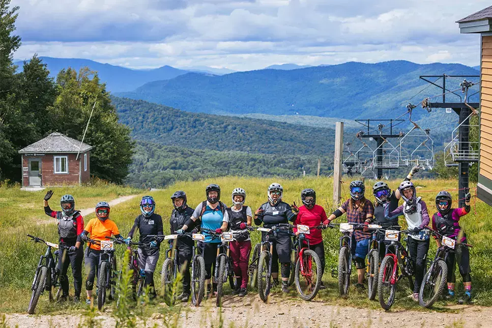 Group of mountain bikers posing on a grassy slope with mountains in the background.