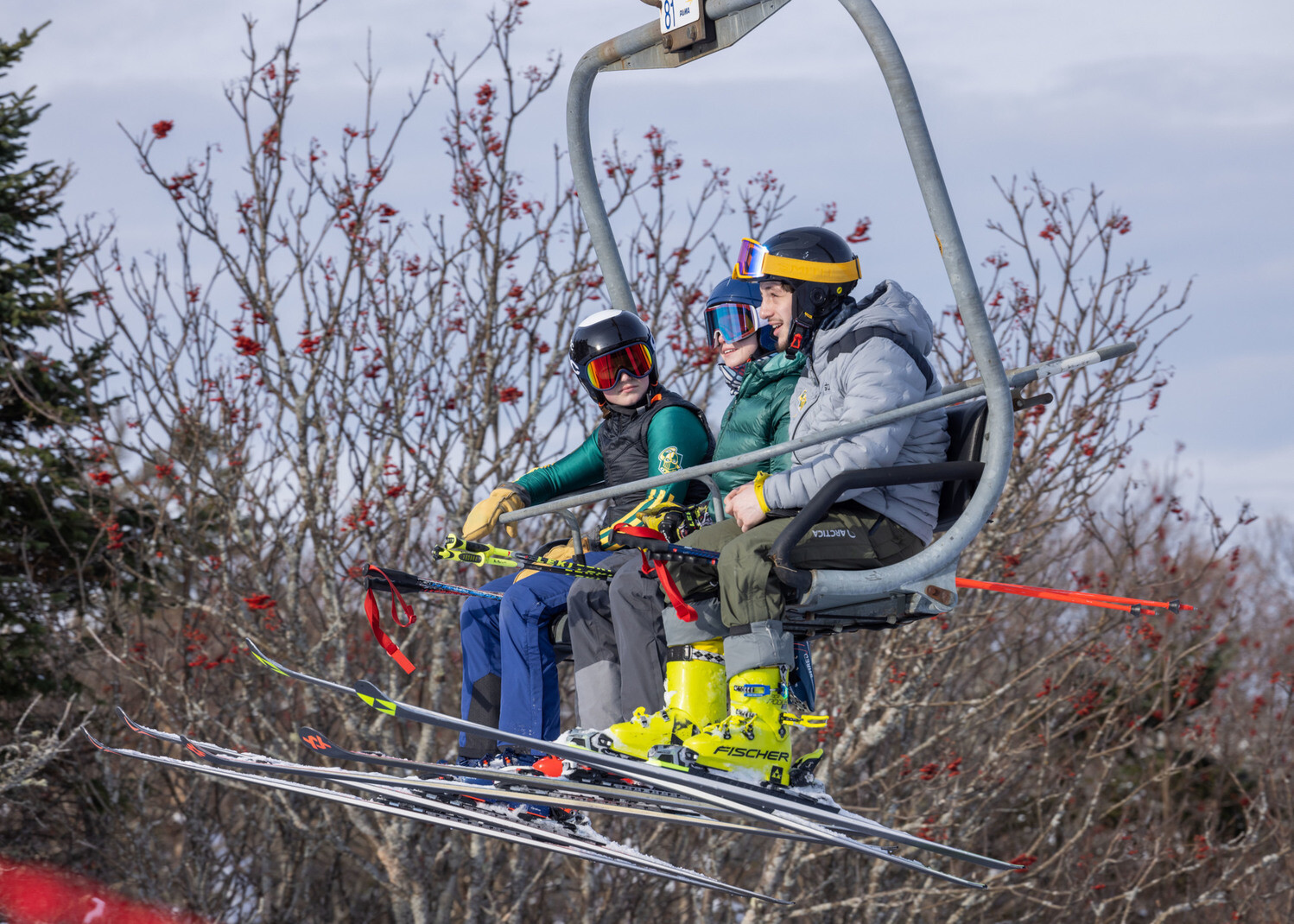 Three skiers on a chairlift, surrounded by trees and a cloudy sky.