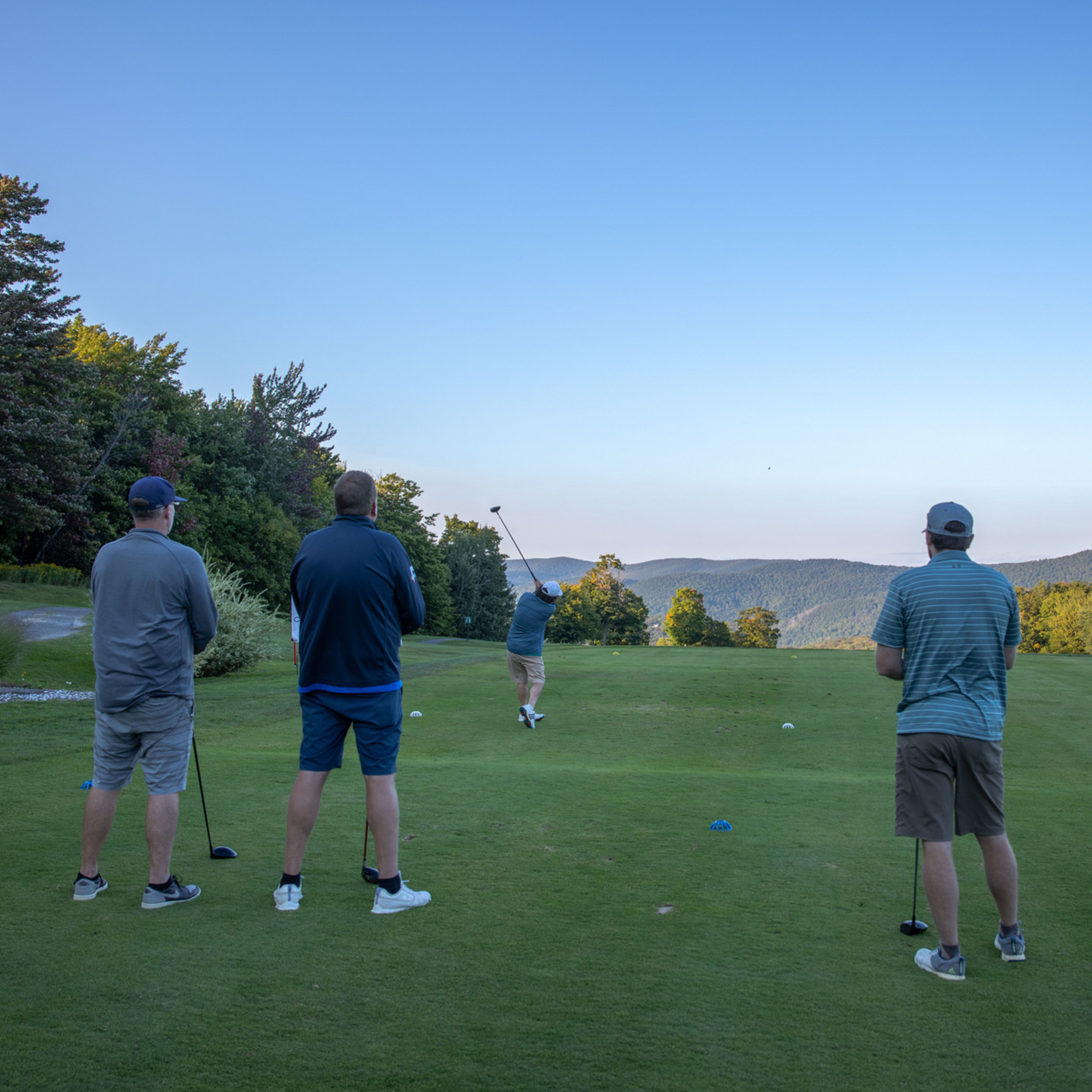 Four men watch as one golfer takes a swing on a scenic golf course.