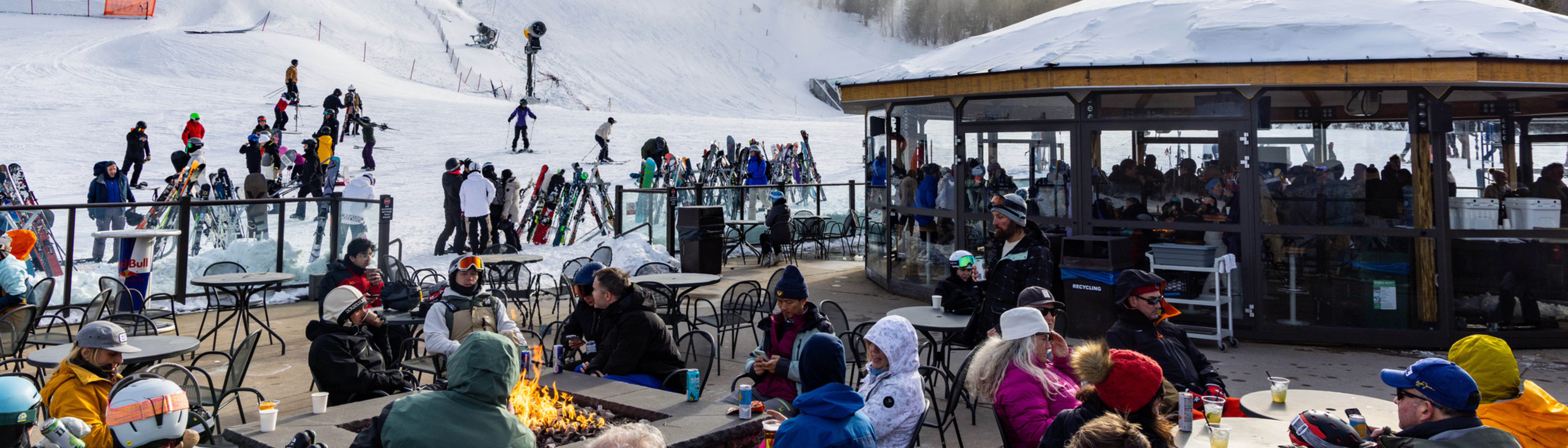 Skiers and snowboarders gather outside a lodge with outdoor seating and a fire pit.