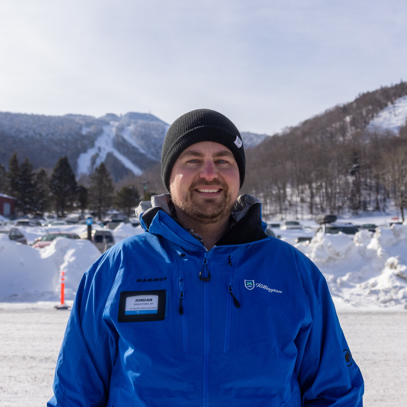 Man in a blue jacket and black beanie, snowy mountains and vehicles in the background.