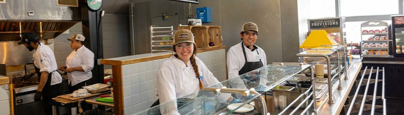 Chefs and staff smile behind a service counter in a busy kitchen setting.