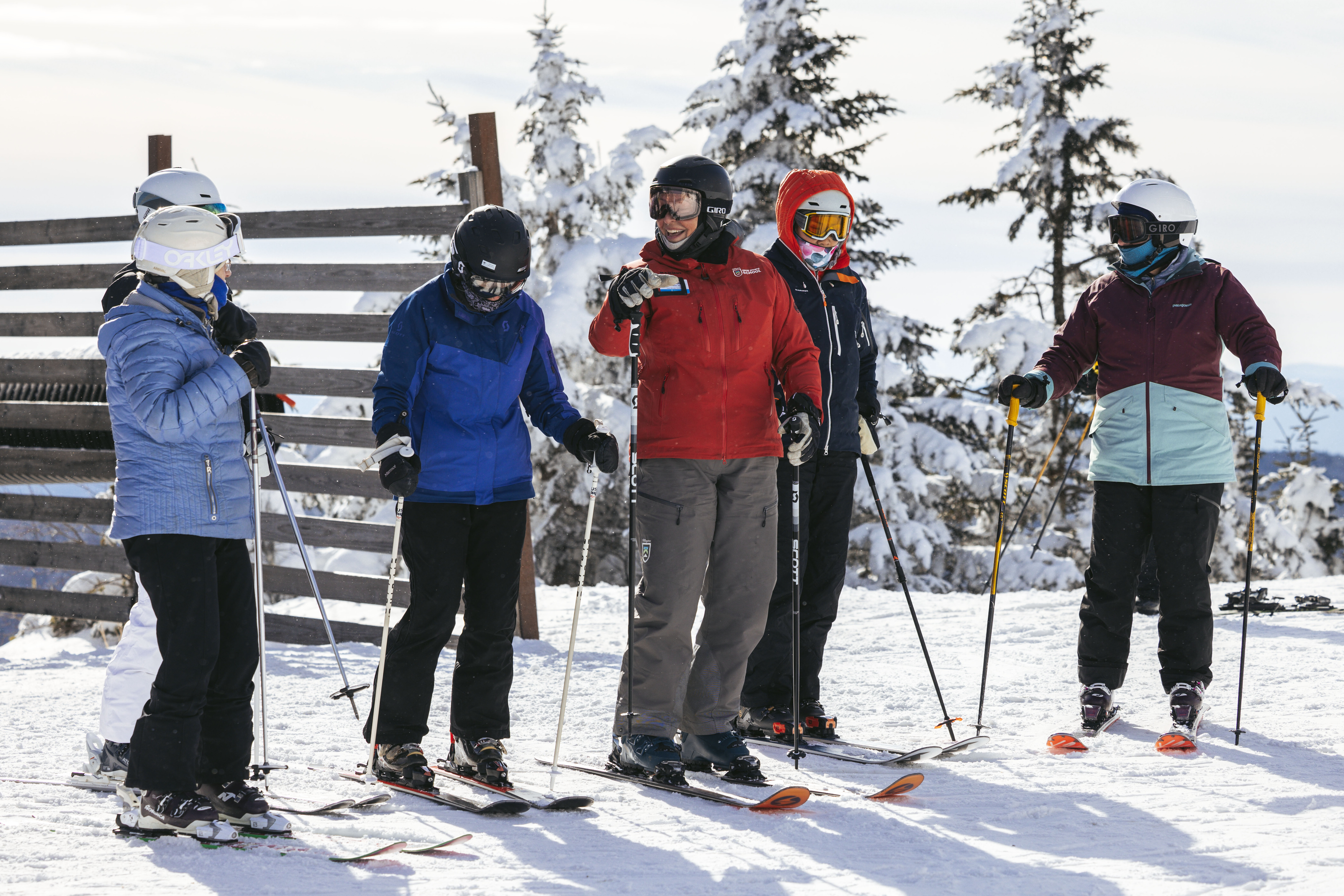 A group of five skiers on a snowy slope, listening to an instructor.
