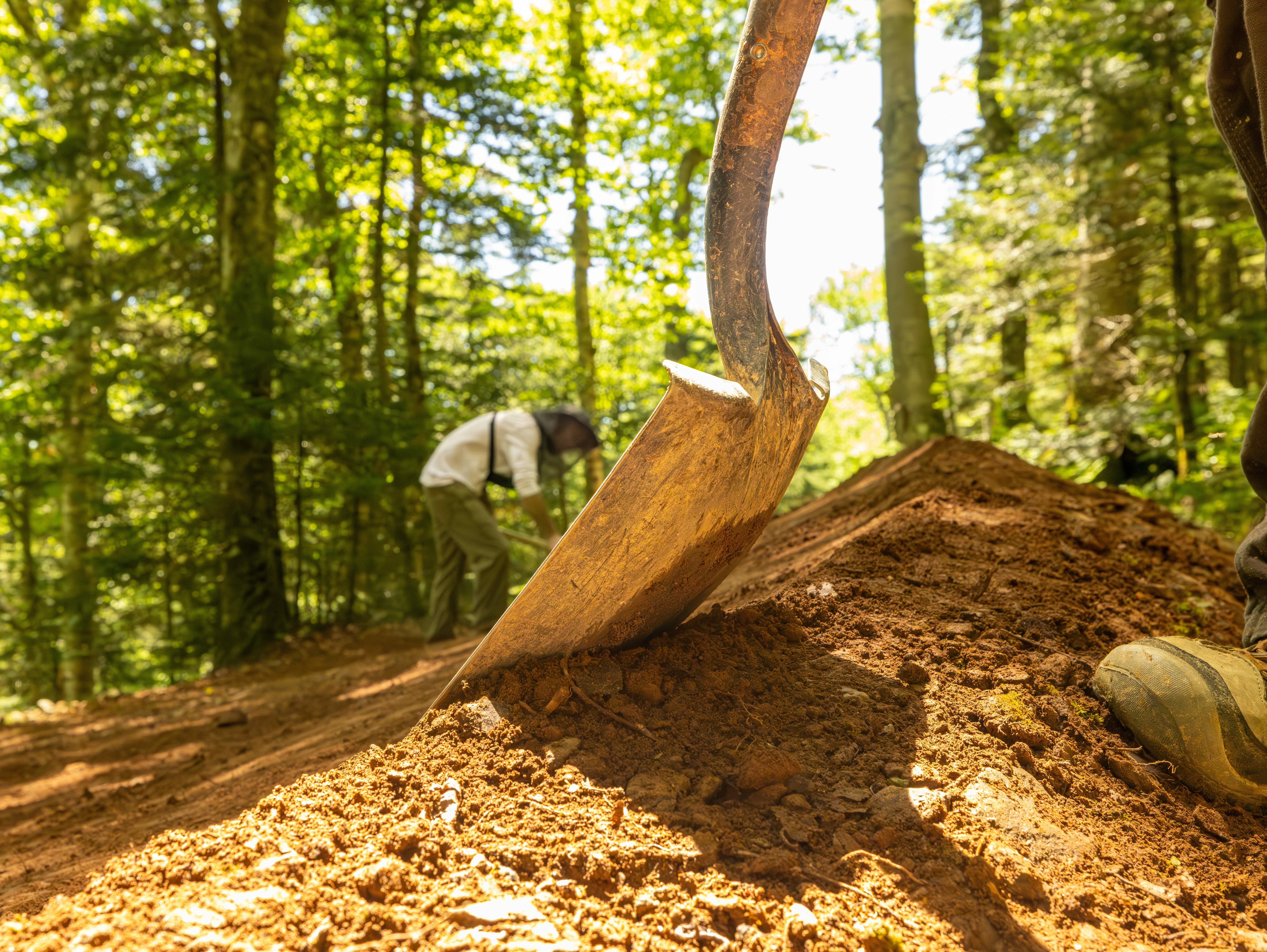 A close-up of a shovel over freshly dug earth, with a person working in the background.