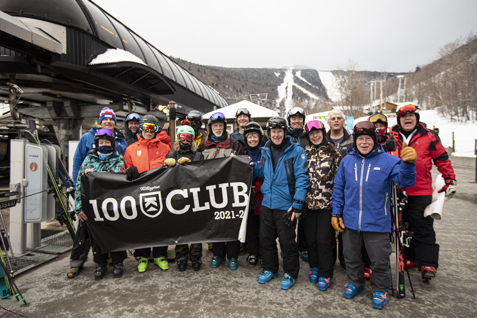 Group of skiers posing with a "100 Club" banner at a ski resort by a chairlift.