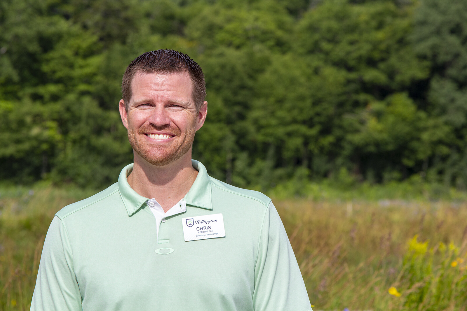 Smiling man in a green polo shirt stands outdoors in a grassy field.