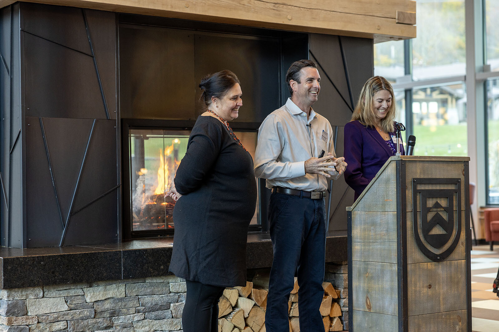 Three speakers at a podium near a fireplace in a modern indoor setting.