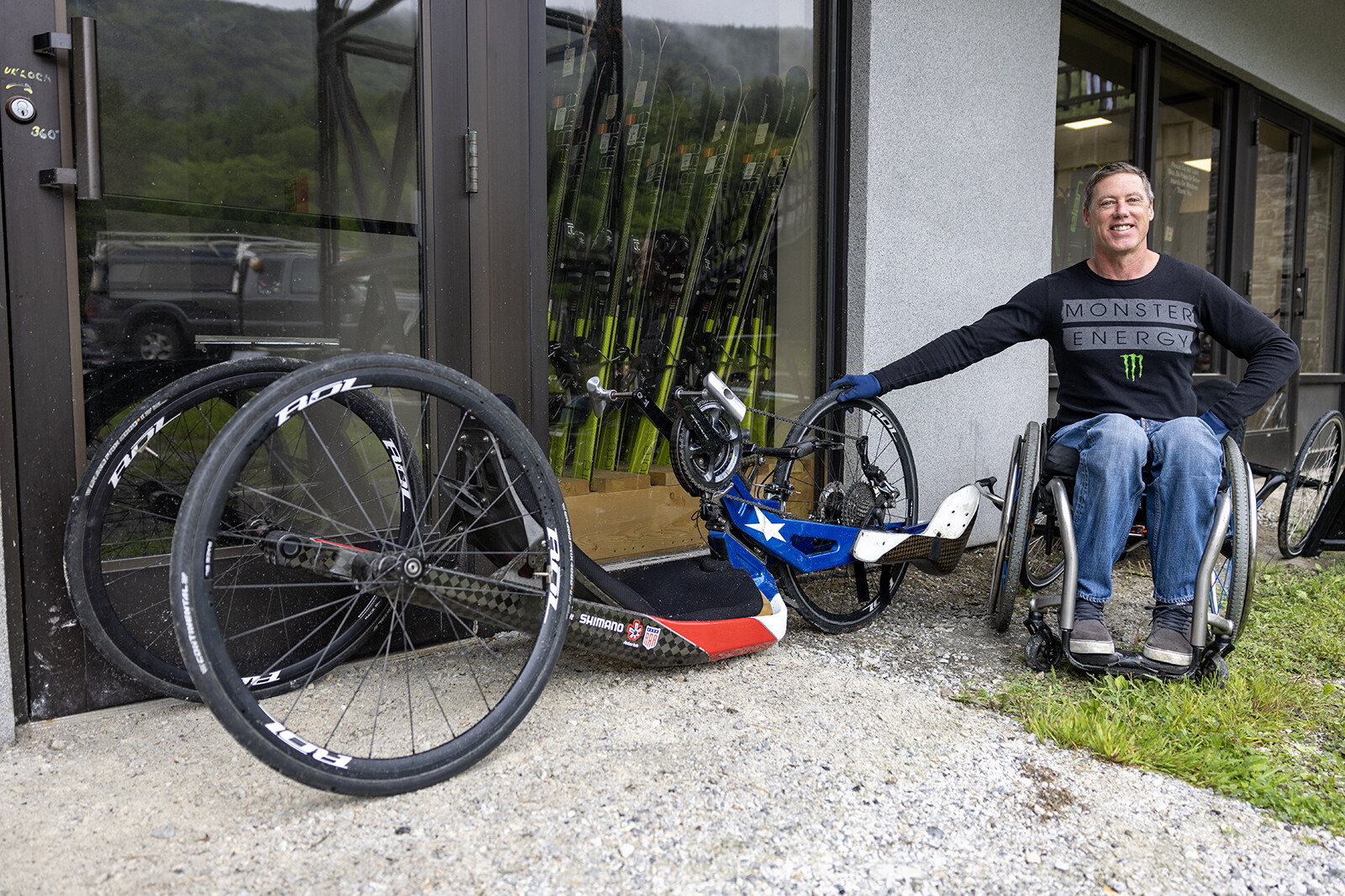 A smiling man in a wheelchair next to handcycles outside a bike shop.
