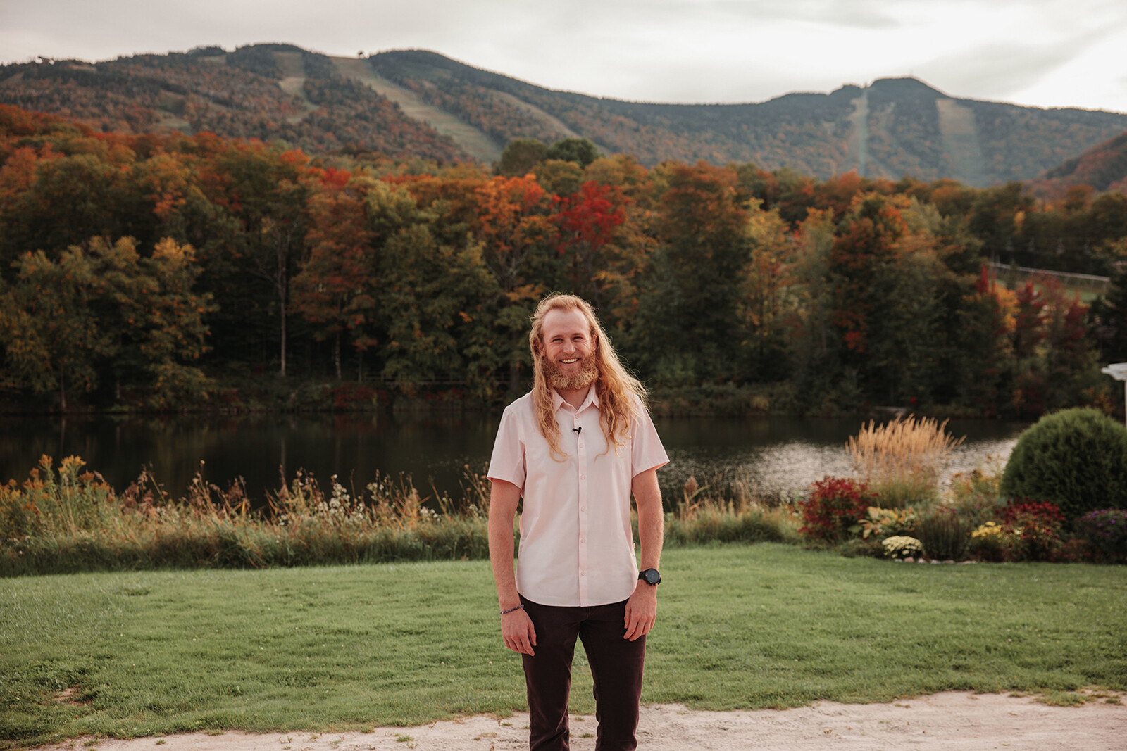 Smiling man with long hair stands by a lake surrounded by colorful autumn trees and mountains.