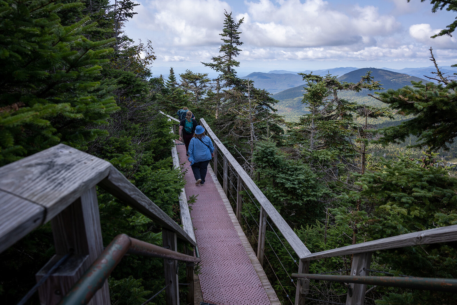 Two hikers walk along a wooden bridge through a forested mountain landscape.