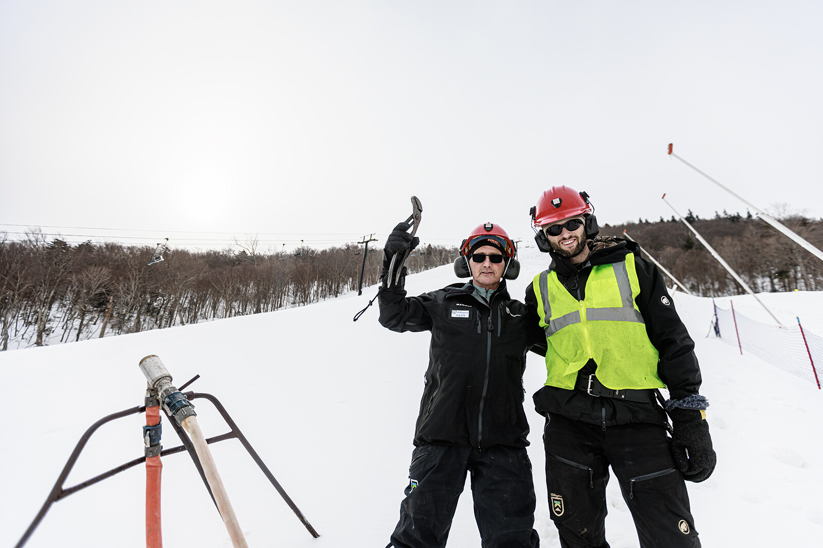 Two ski workers pose in snow, wearing helmets and gear, with ski lift in background.