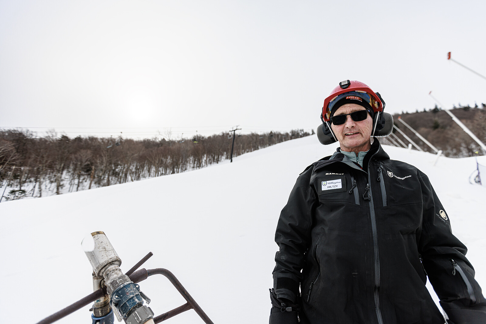 A ski resort worker stands in winter attire on a snowy slope with trees in the background.