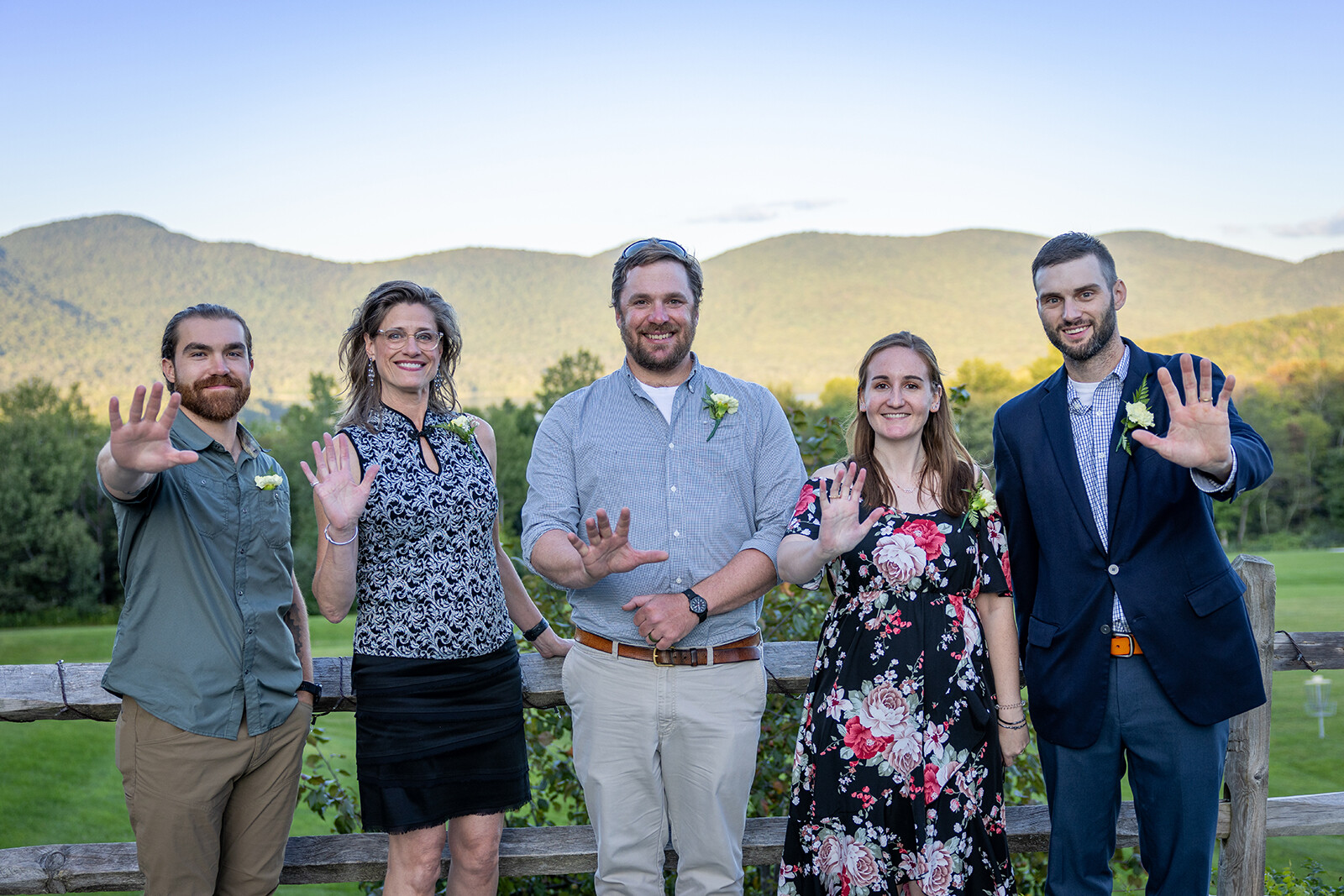Five smiling individuals wave against a scenic mountain backdrop.