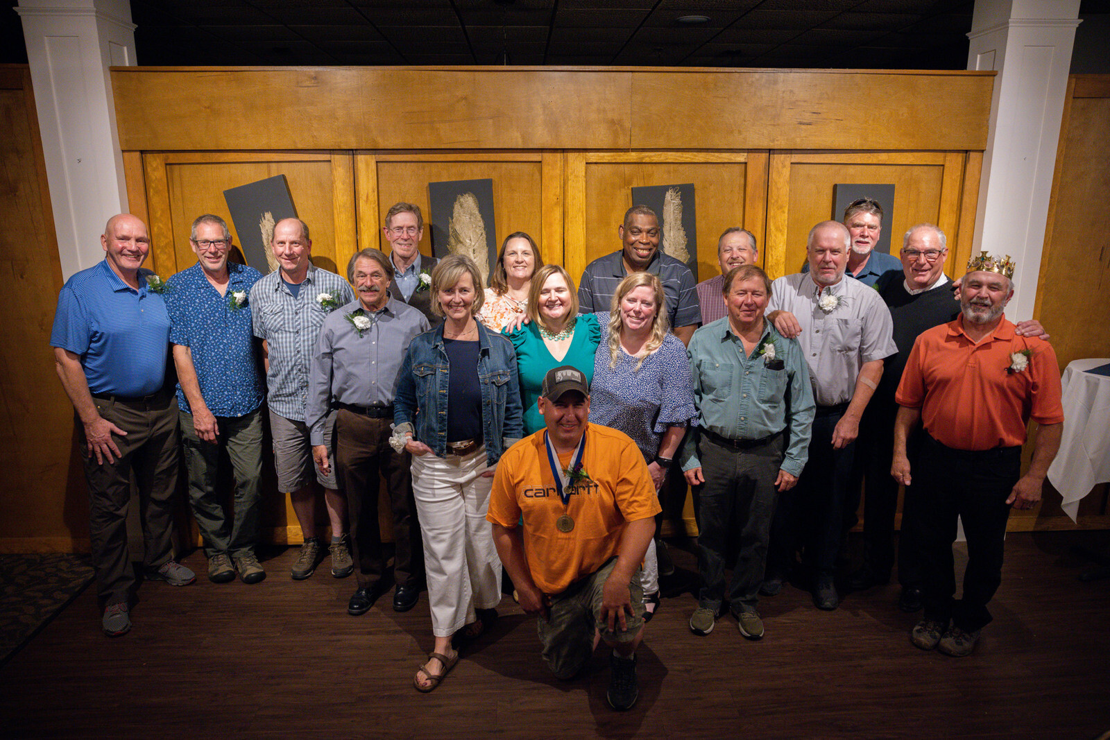 Group photo of 16 people posing indoors, smiling, with floral decorations on their outfits.