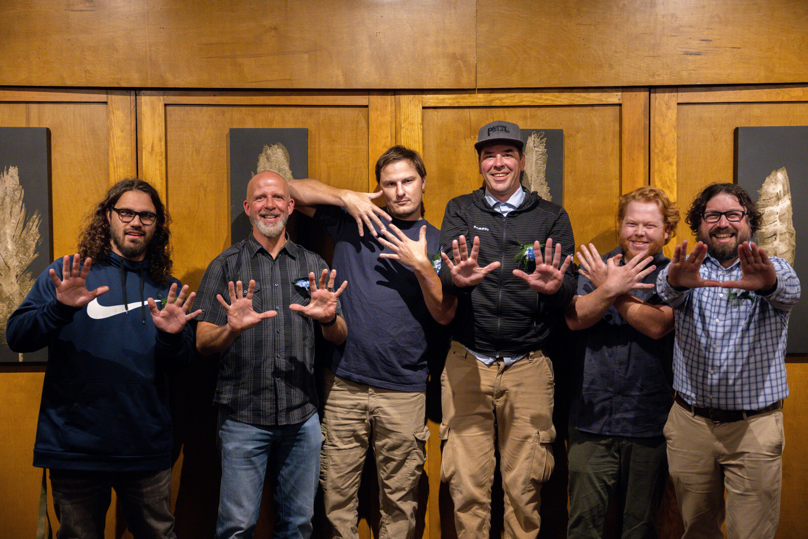 Six people pose together, raising hands in a playful manner against a wooden backdrop.