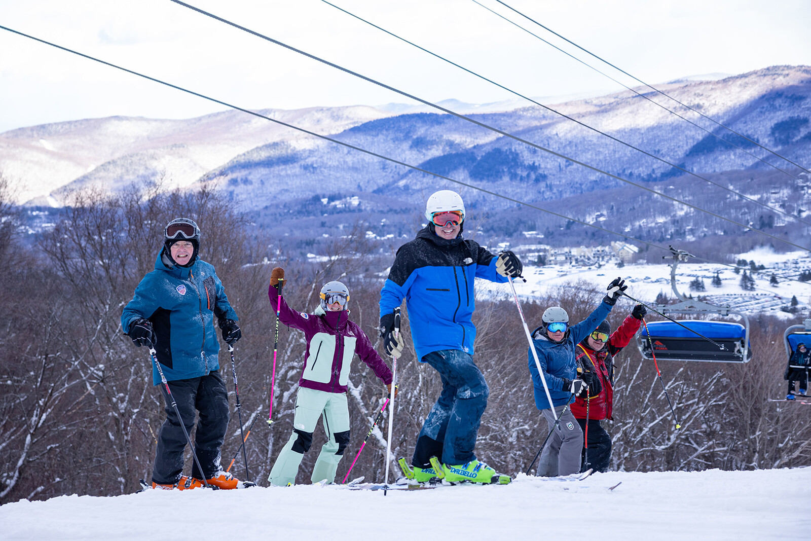 Five skiers on a snowy slope, smiling, with mountains and ski lifts in the background.