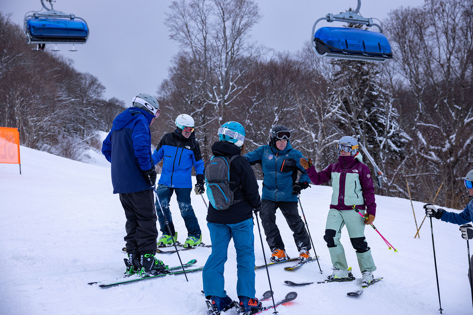 A group of six skiers gather on a snowy slope, chatting under a ski lift.