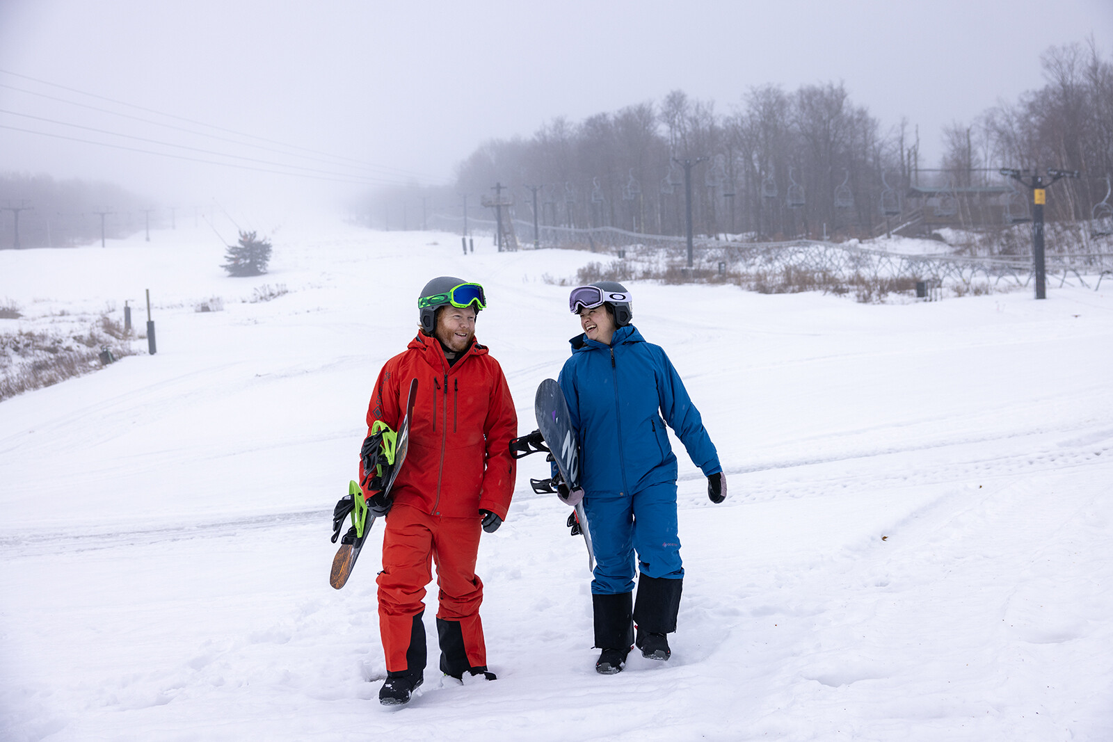 Two snowboarders in winter gear walk together on a snowy hill.