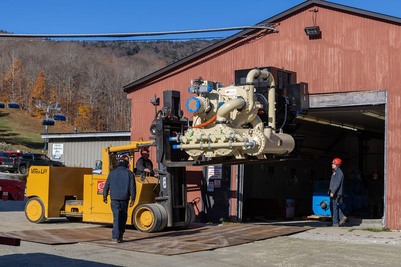 Forklift lifting equipment outside a red barn with workers nearby. Fall foliage visible.