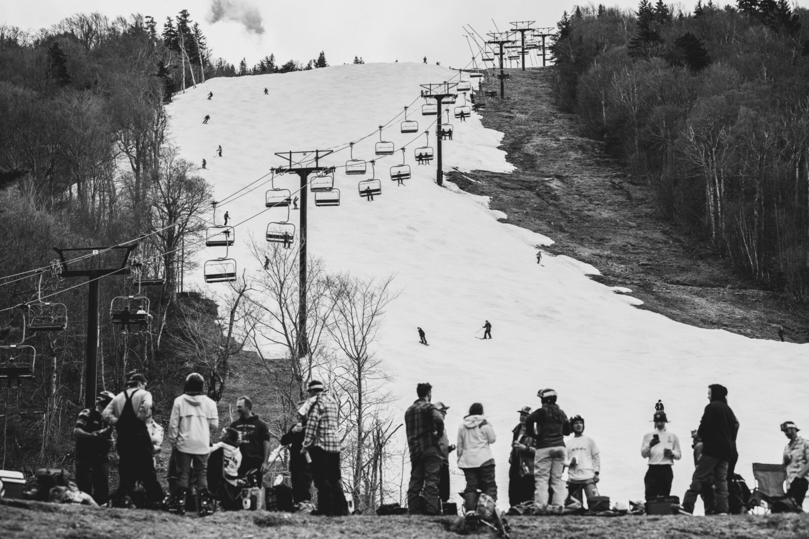Skiers on a snowy slope with a lift, while a group gathers below. Black and white photo.