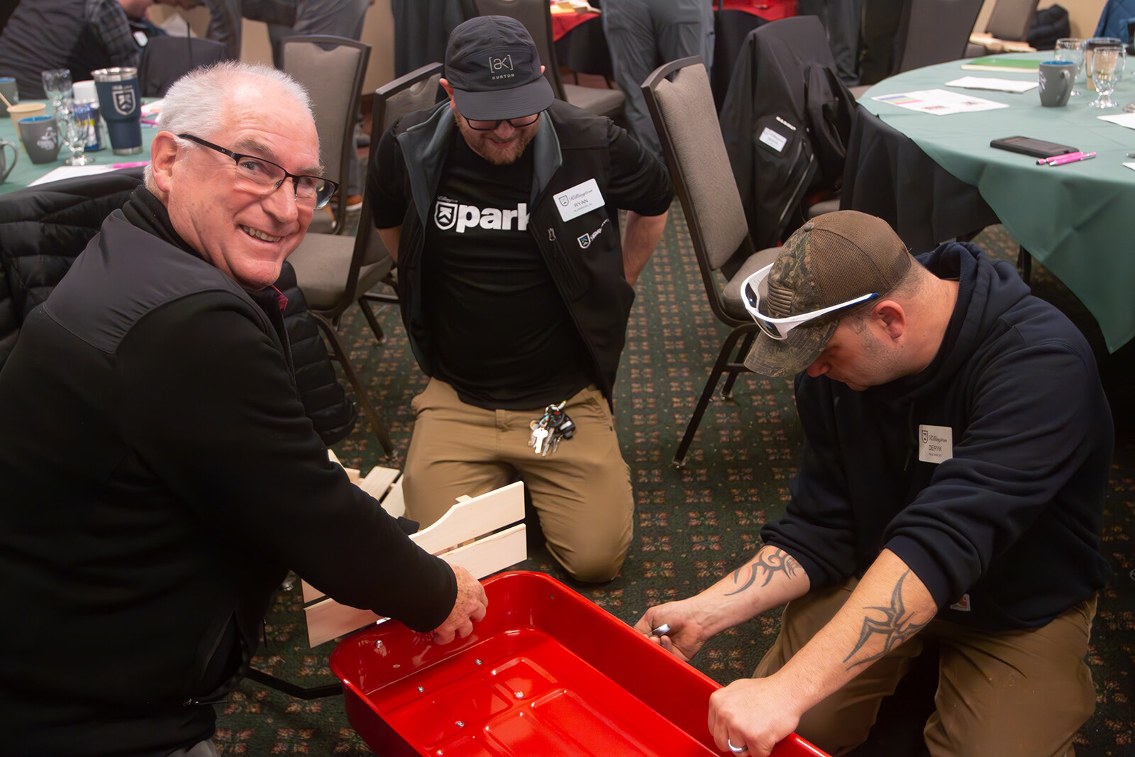 Three men work together building a red wagon in a conference room setting.