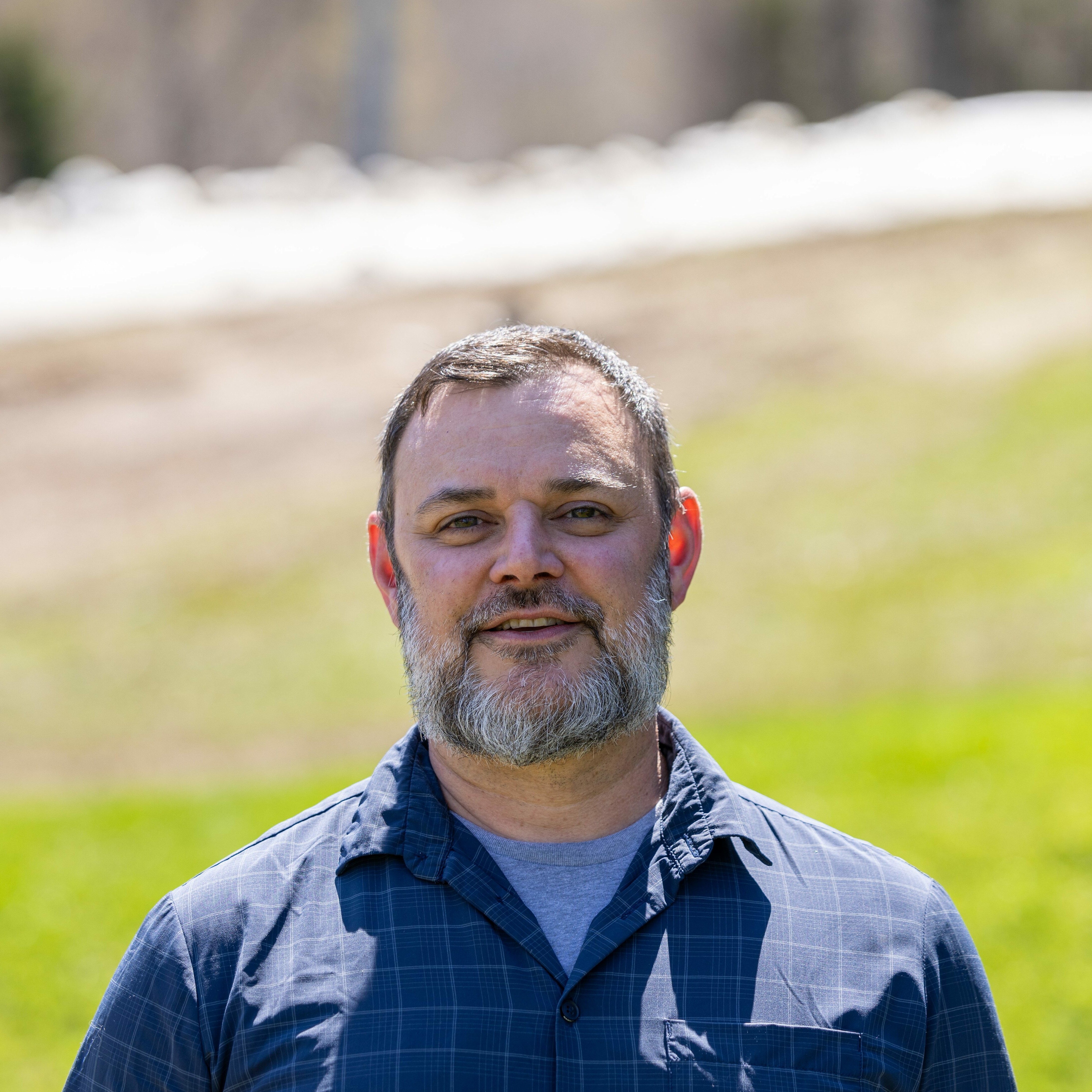 Smiling man with a beard stands outdoors against a blurred green and brown background.