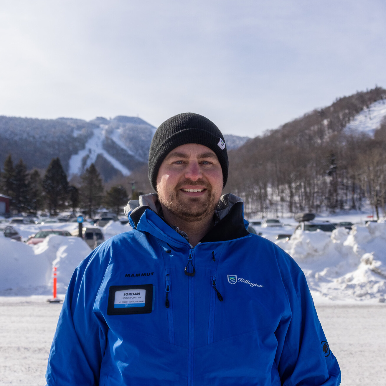 Man in a blue jacket stands outdoors in snowy mountains, with ski slopes visible behind.