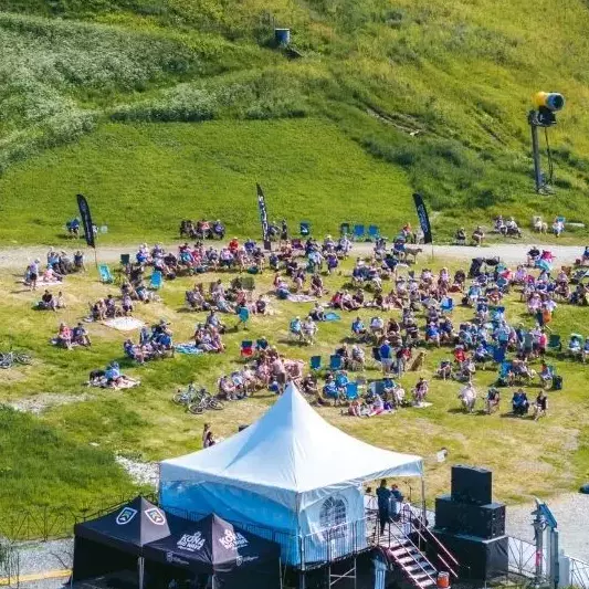 A large crowd sits on grass enjoying an outdoor event near a stage and tents.