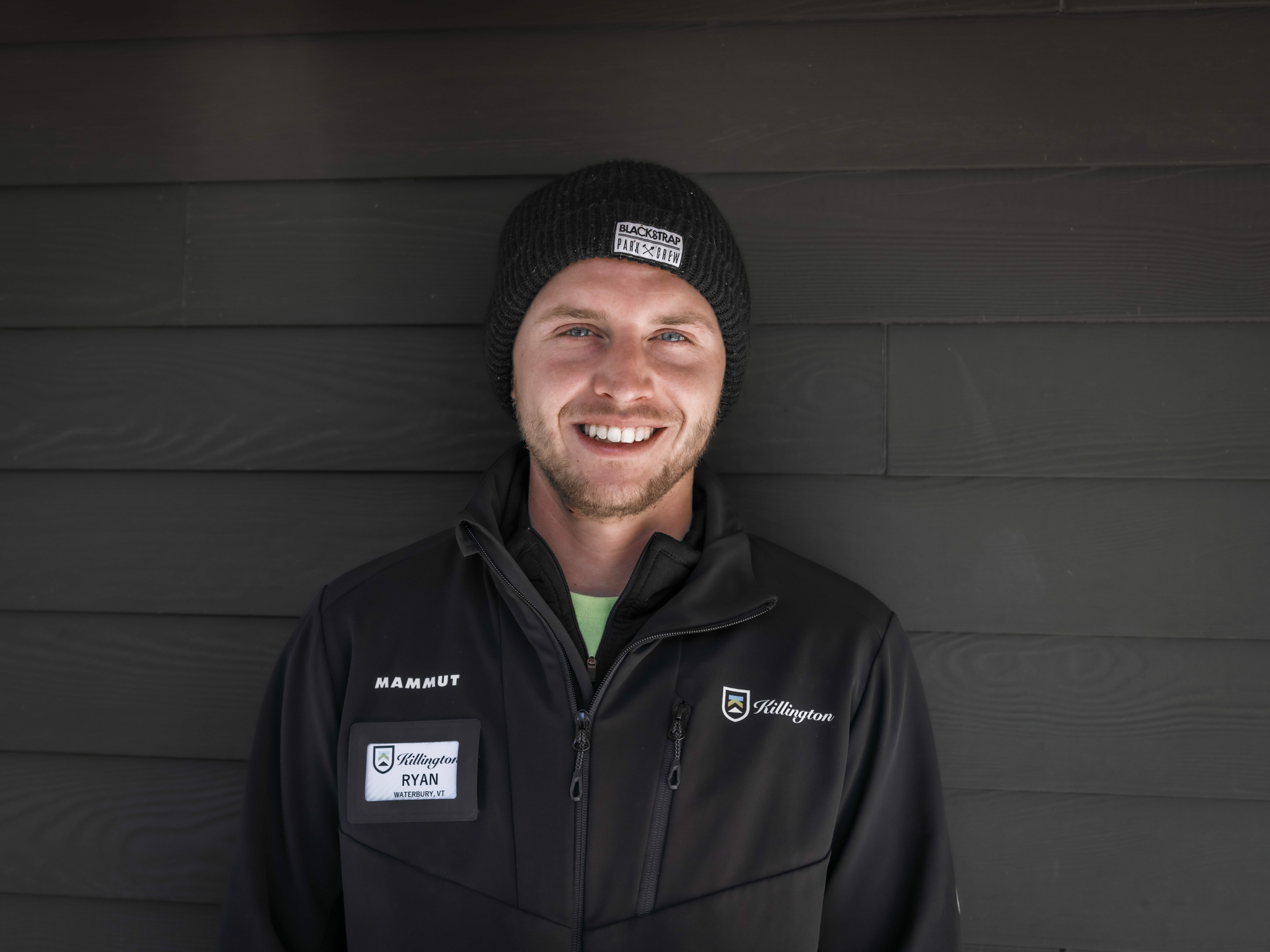 Smiling man in a black jacket and beanie stands against a dark wooden wall.