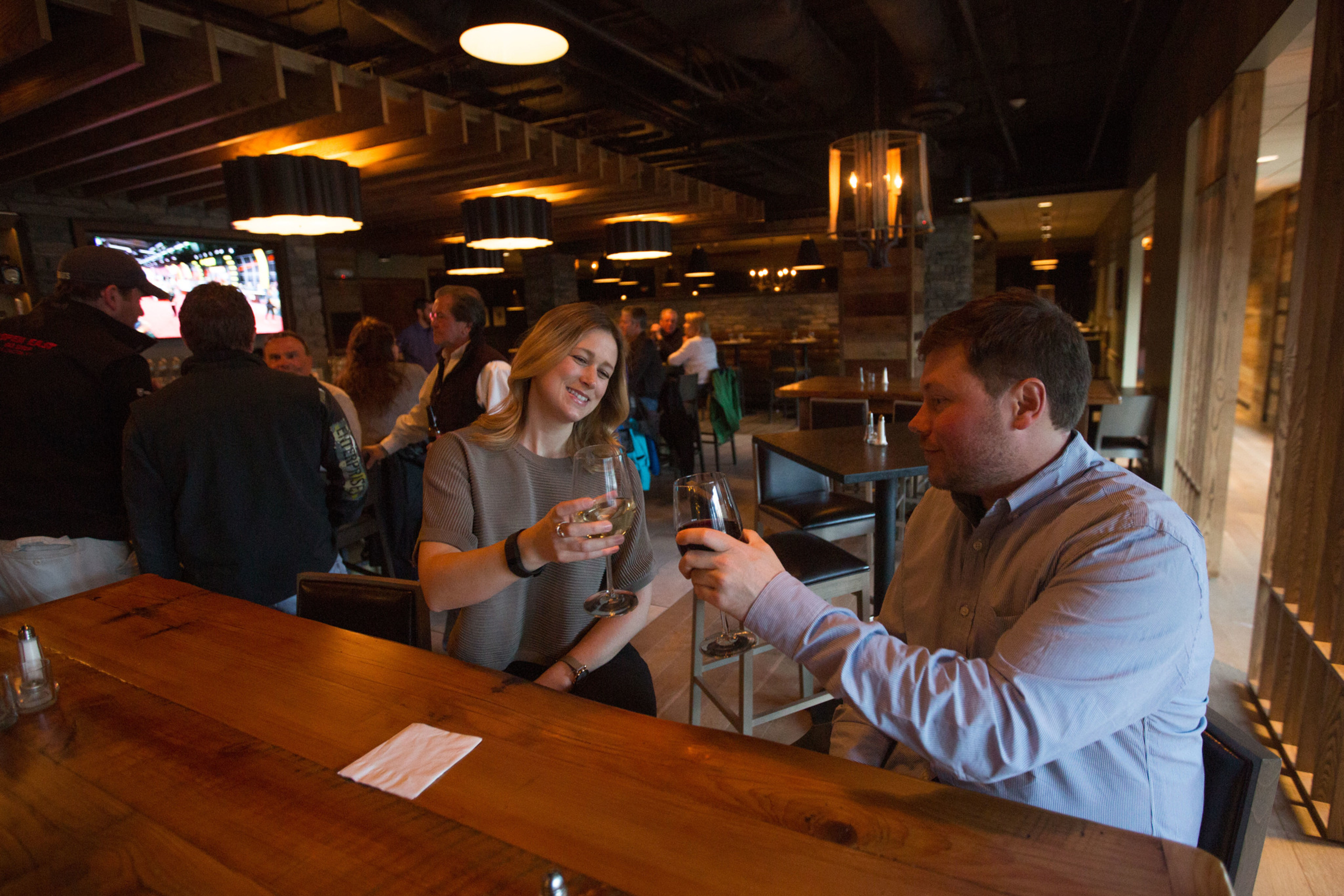 A woman and a man toast glasses of wine at a bar, smiling amidst a lively restaurant atmosphere.